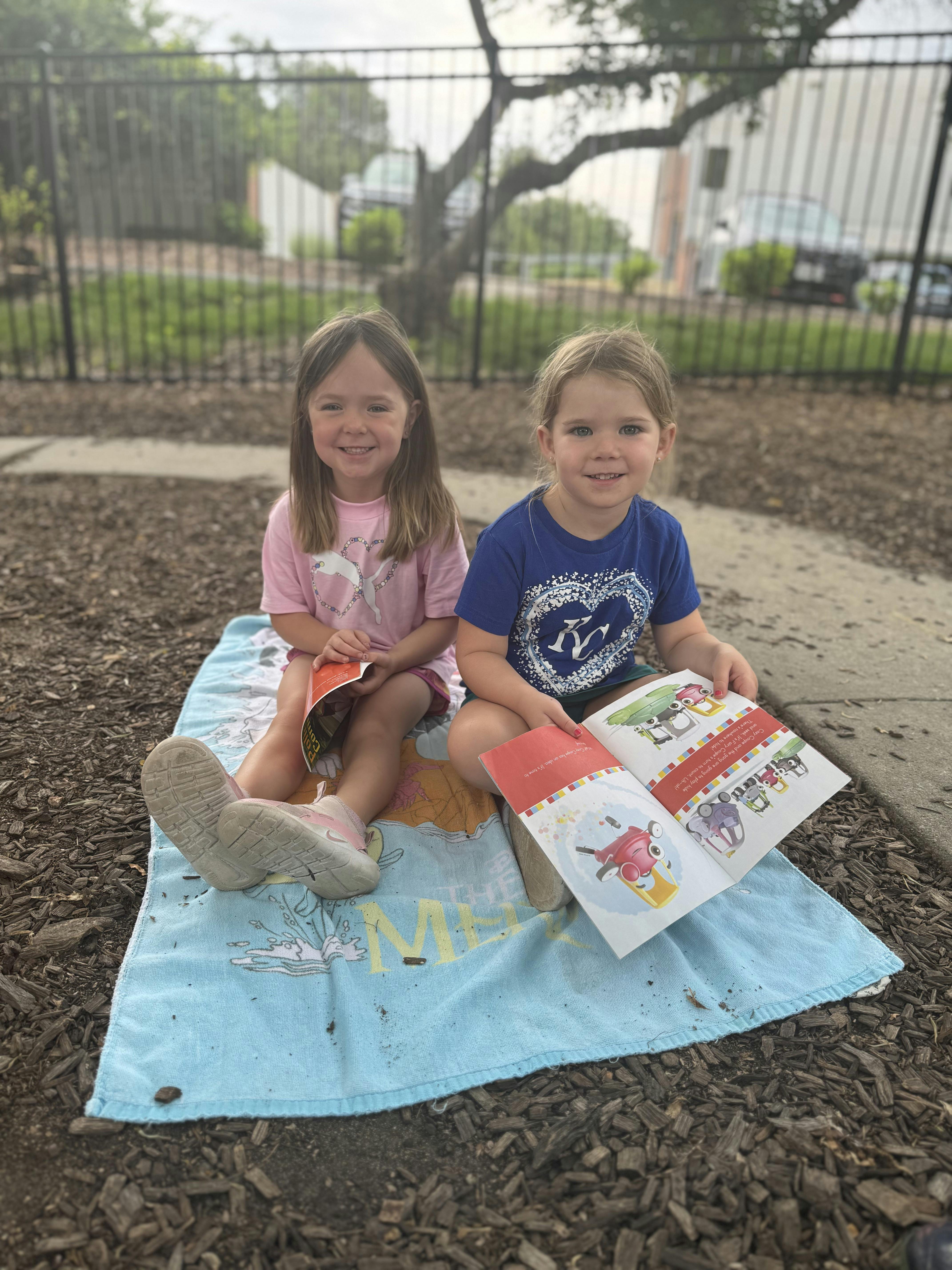 Two children sitting on a towel outdoors reading a book together