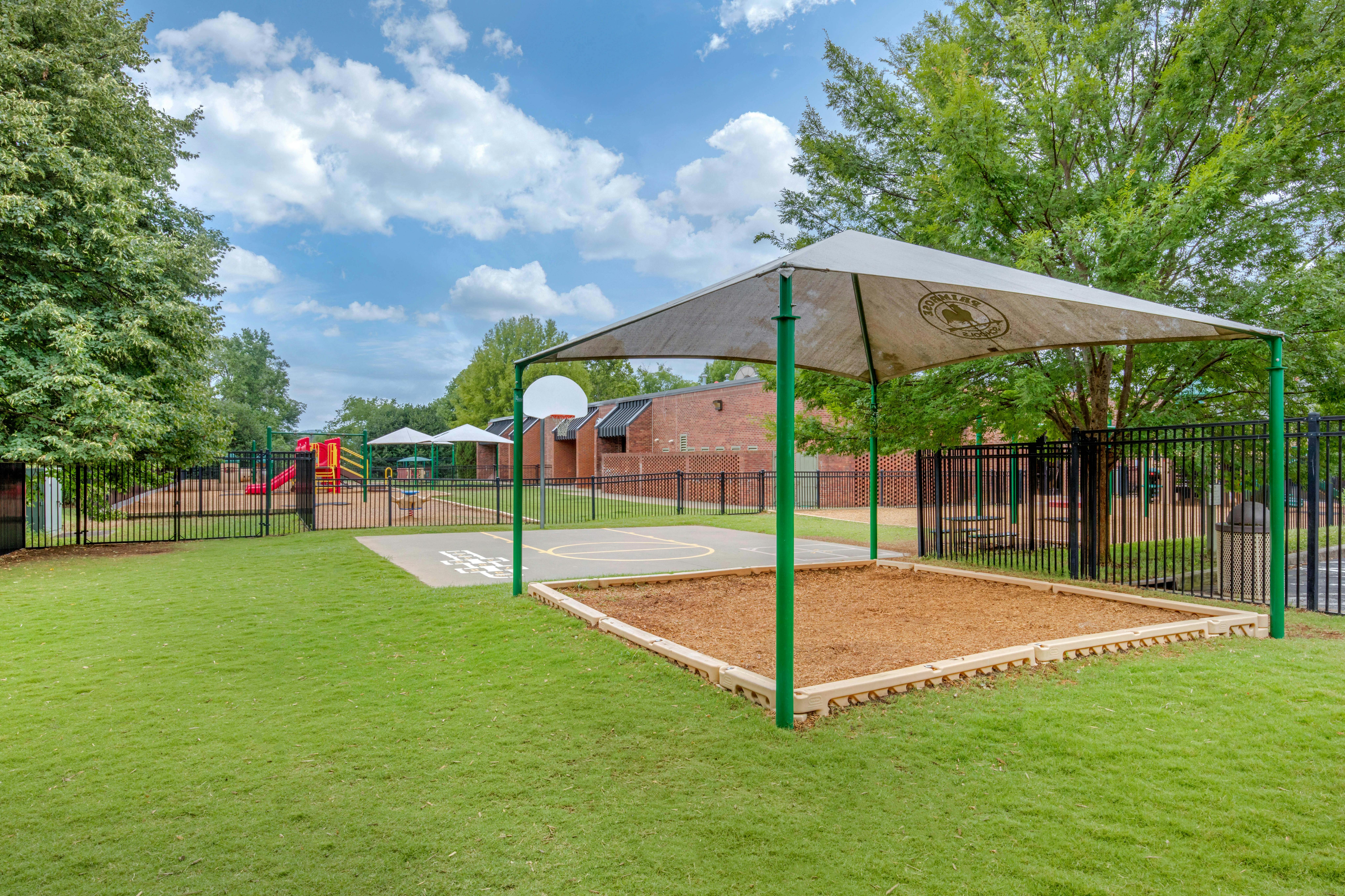 Outdoor playground with a small basketball court and sandpit under a shade structure