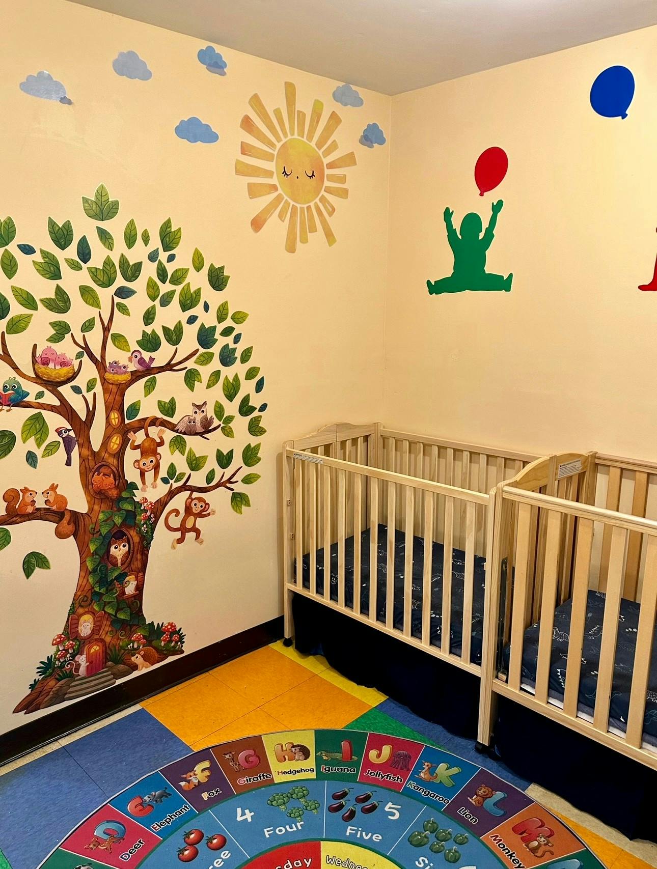 Two cribs in a room with a tree and sun mural on the wall
