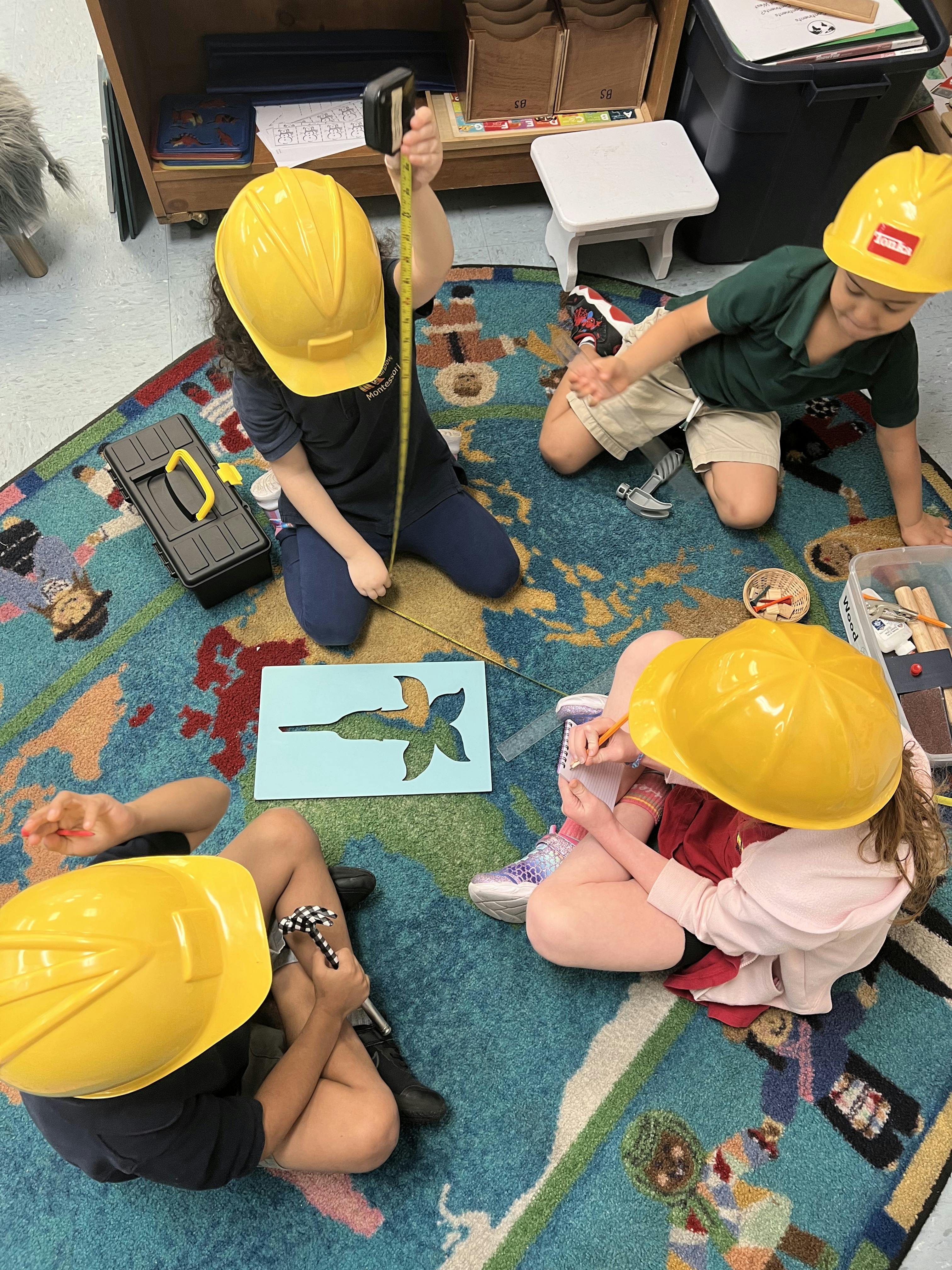 Four children wearing yellow construction hats playing with tools on a rug