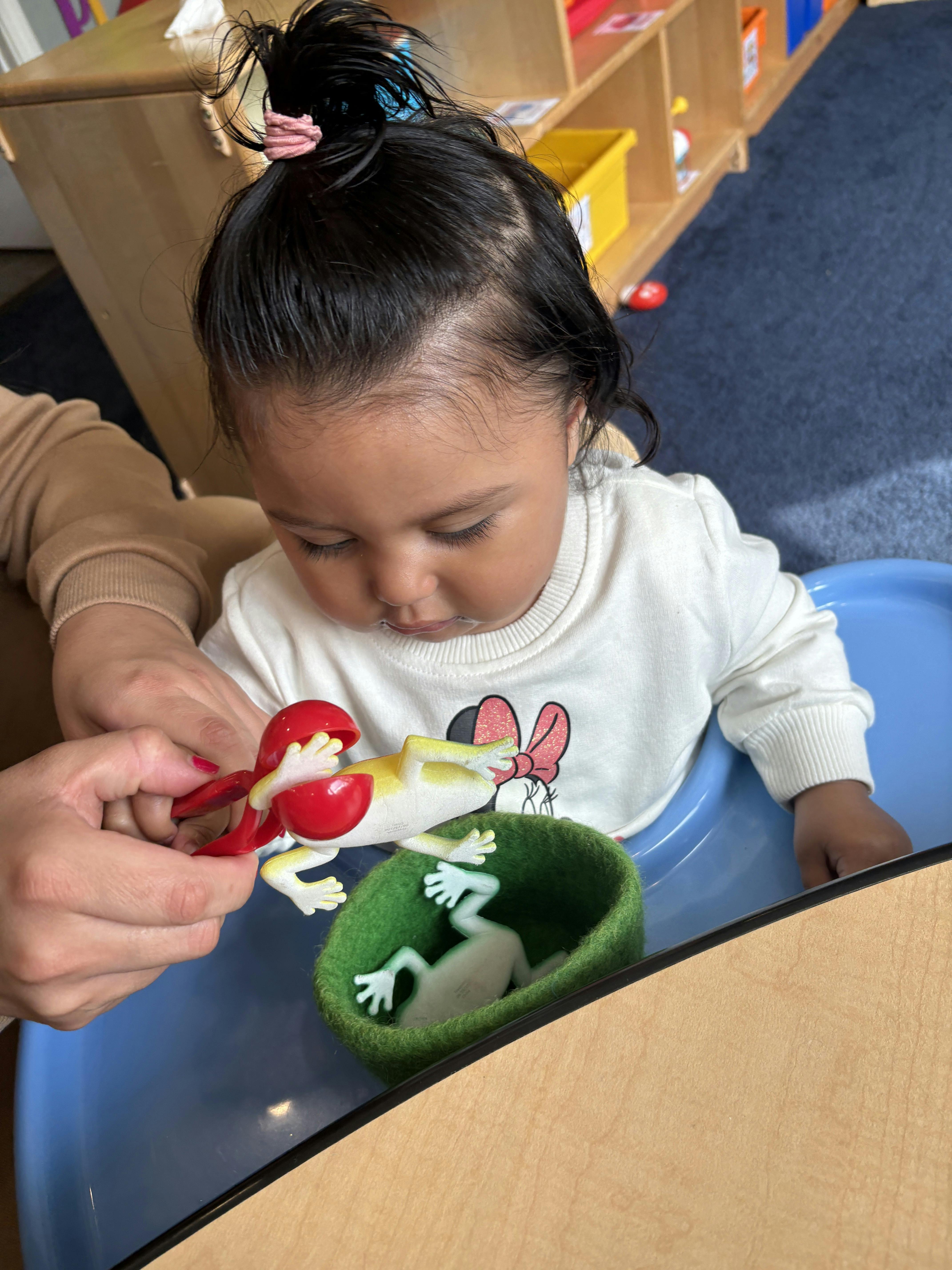 A person helping a child place toy frogs into a small green felt basket