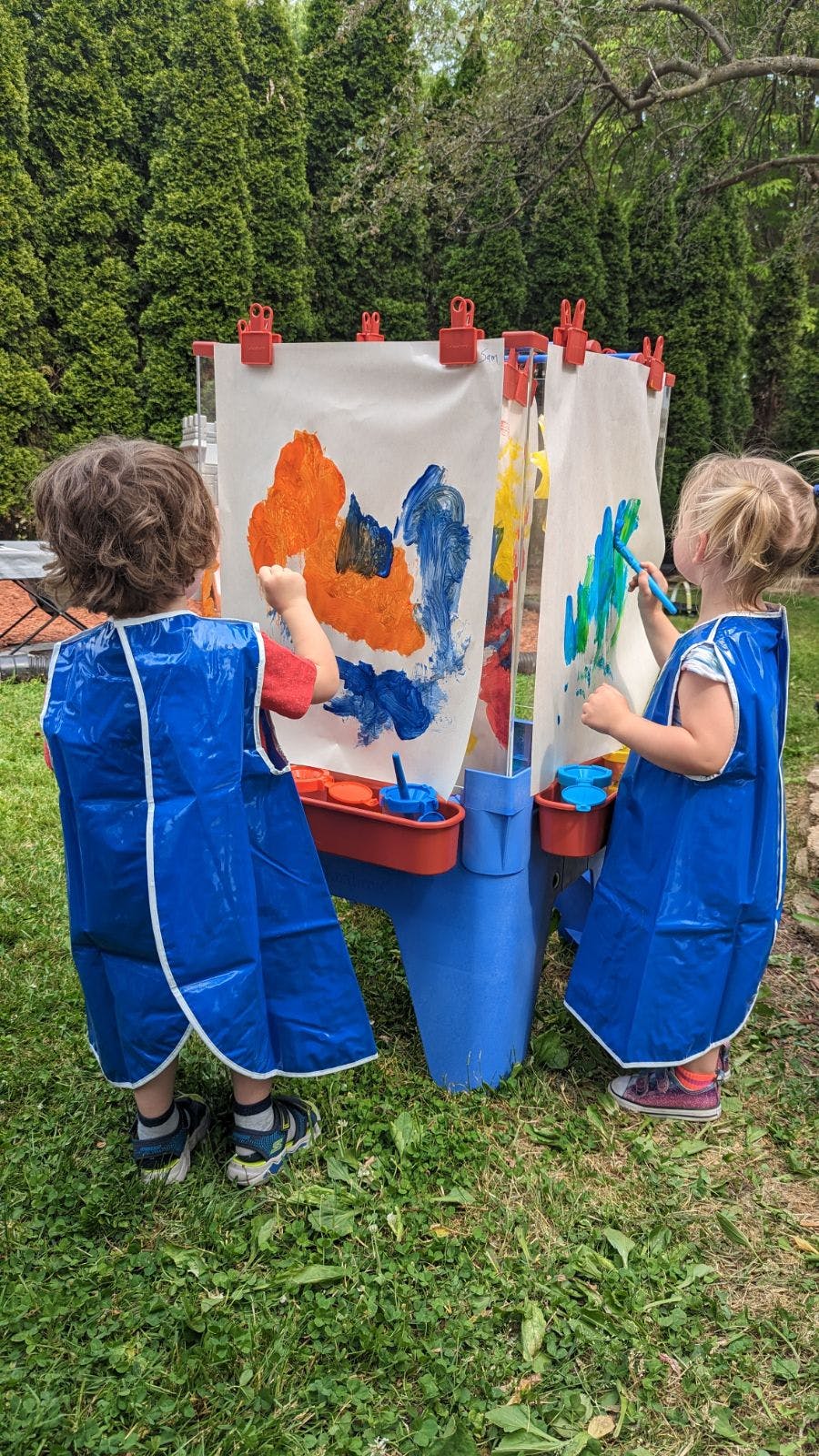 Two children in blue smocks painting at an outdoor easel