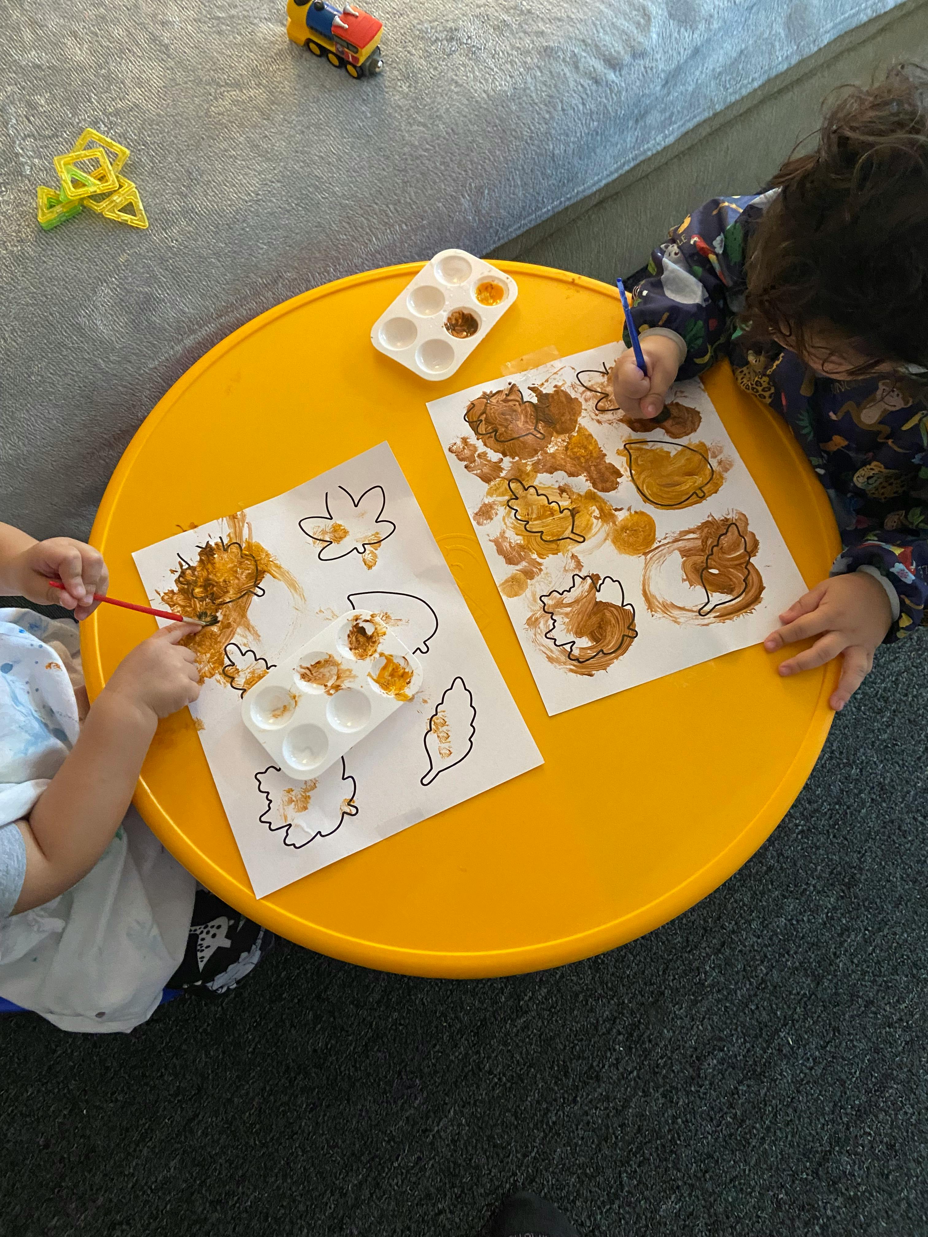 Two children painting leaf outlines on a yellow table