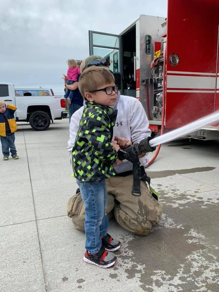 Child in glasses holding a fire hose with a person kneeling behind