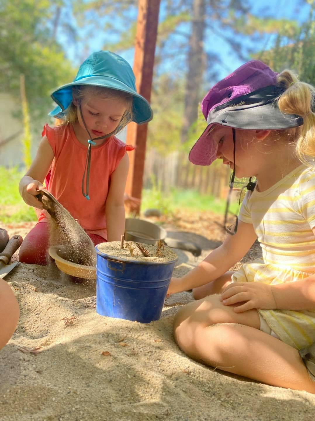 Two children playing in a sandbox with a blue bucket