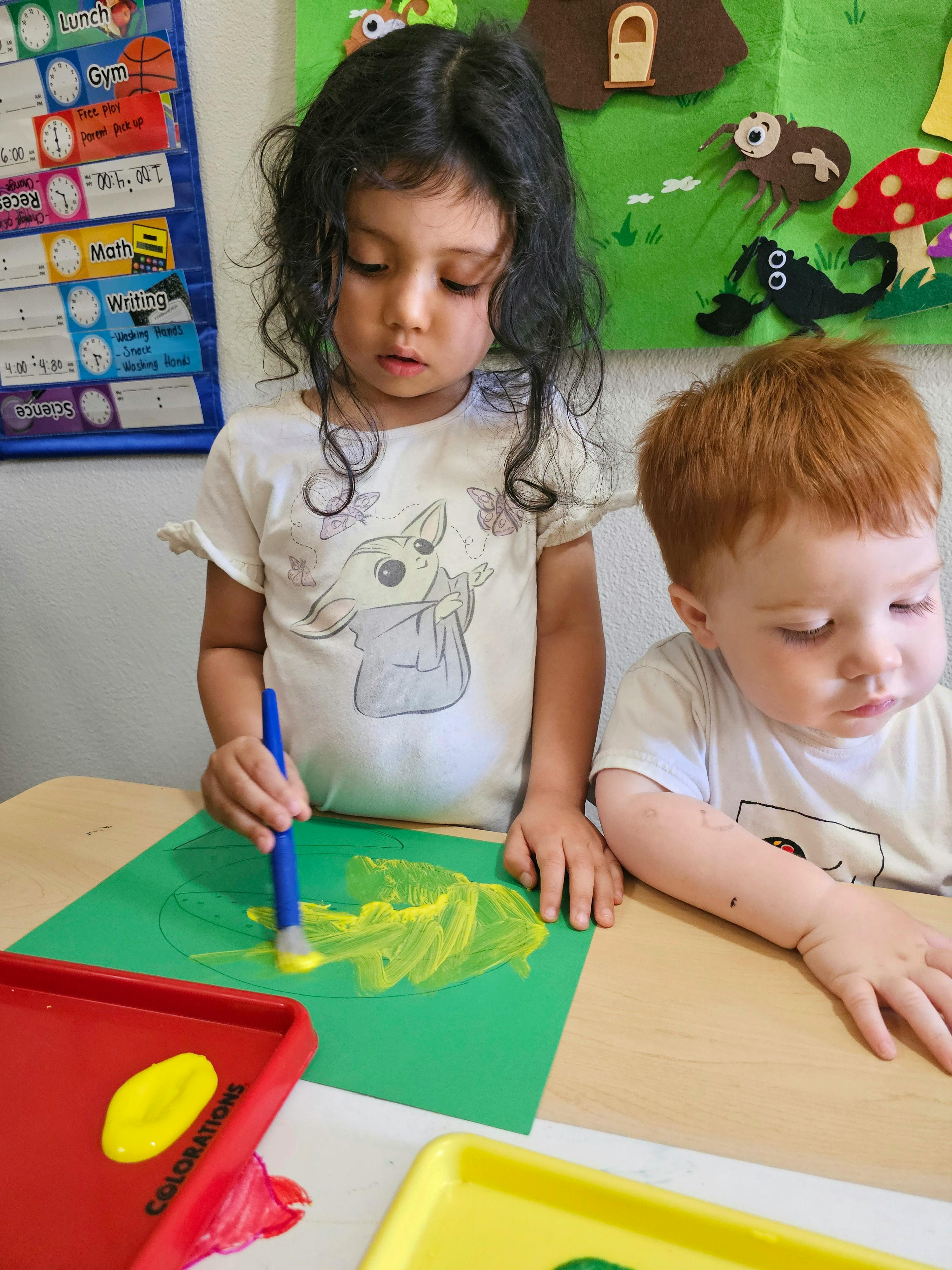 Two children painting on paper at a table in a classroom