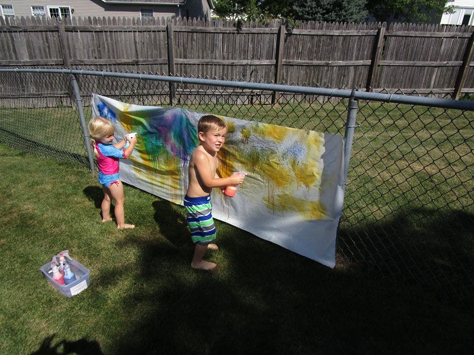 Two children spraying paint onto a white sheet hanging on a fence