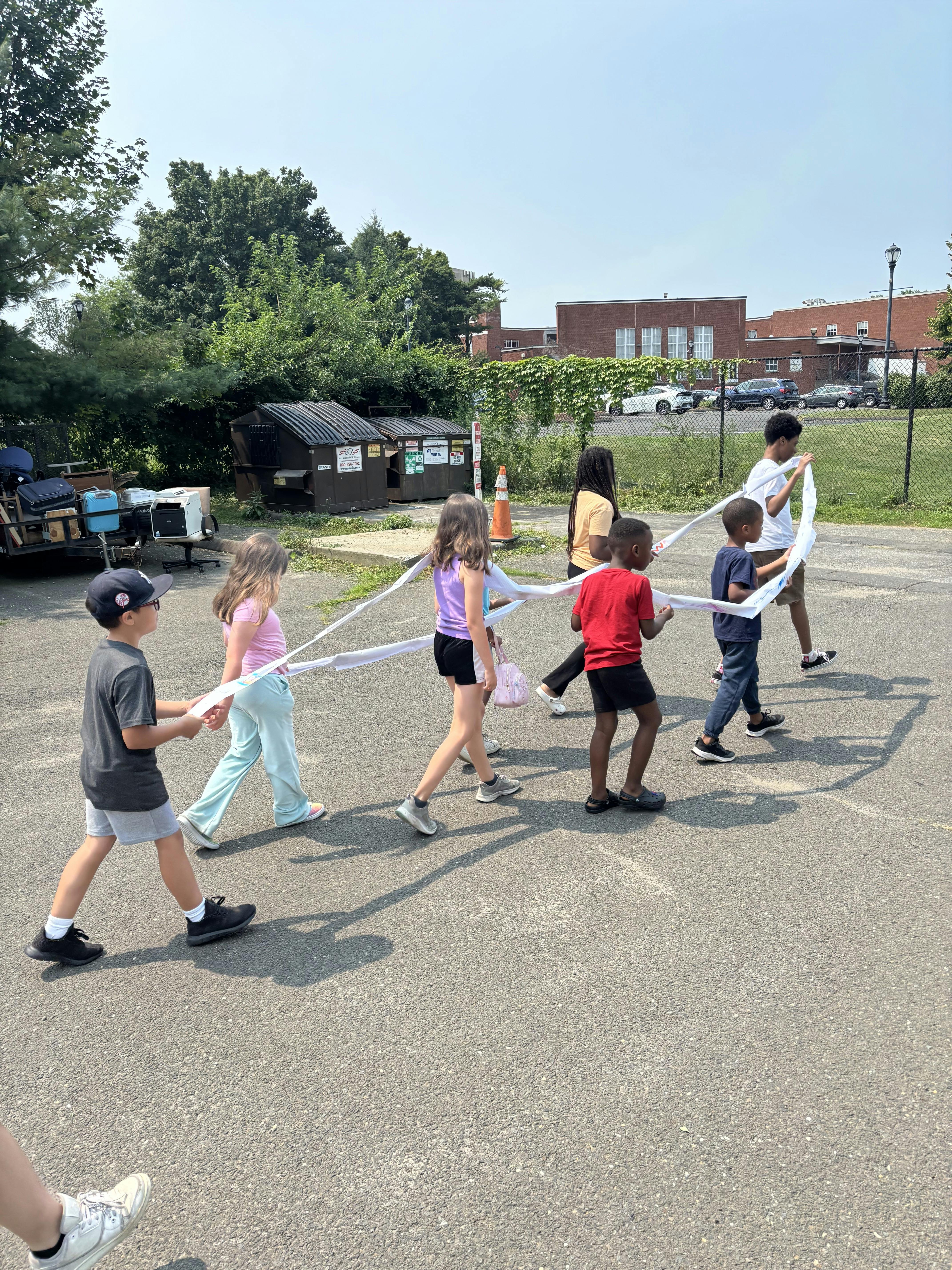 A group of children walking in a line while holding a long strip of white paper