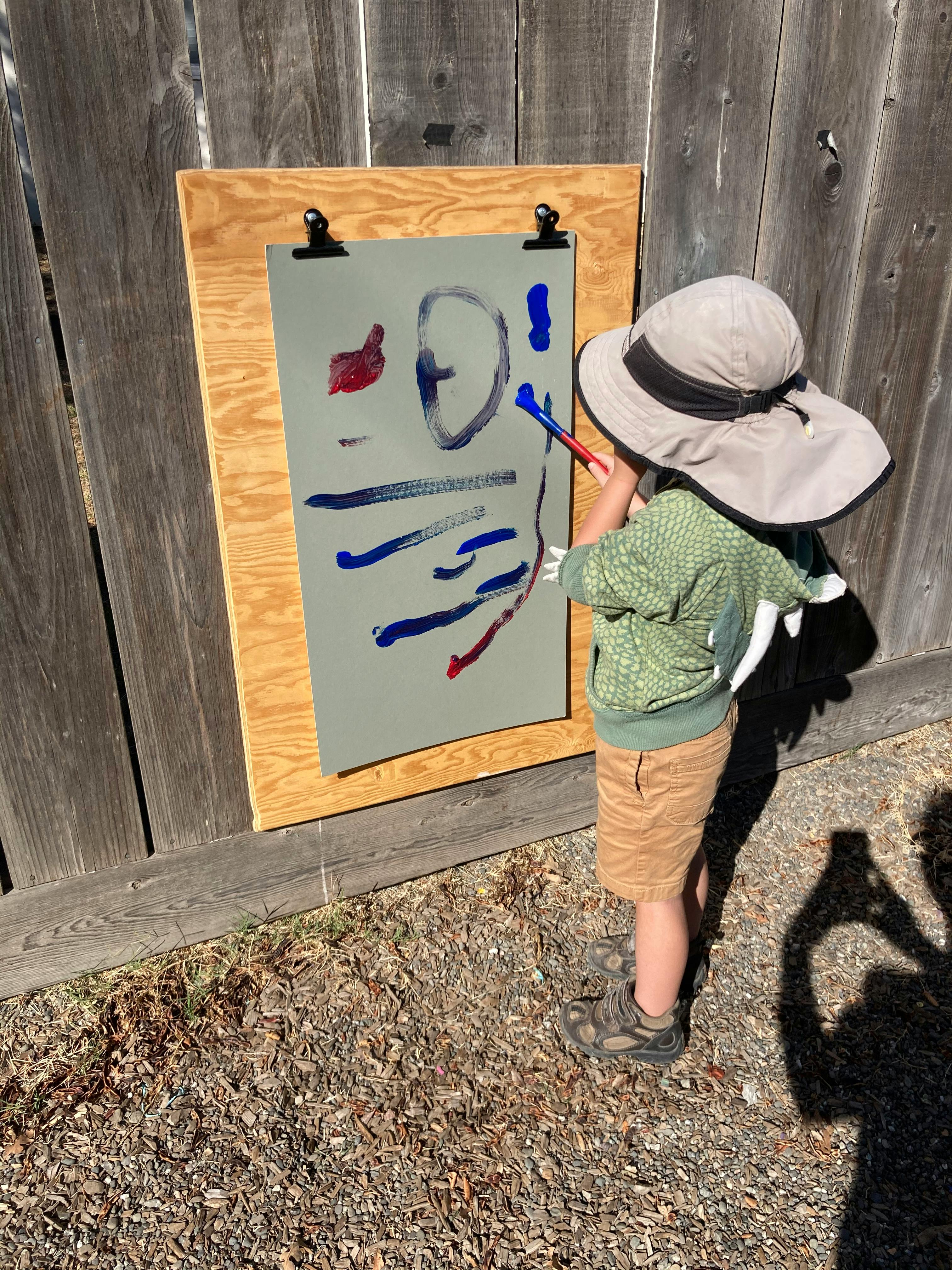 Child in a hat painting on an easel outdoors