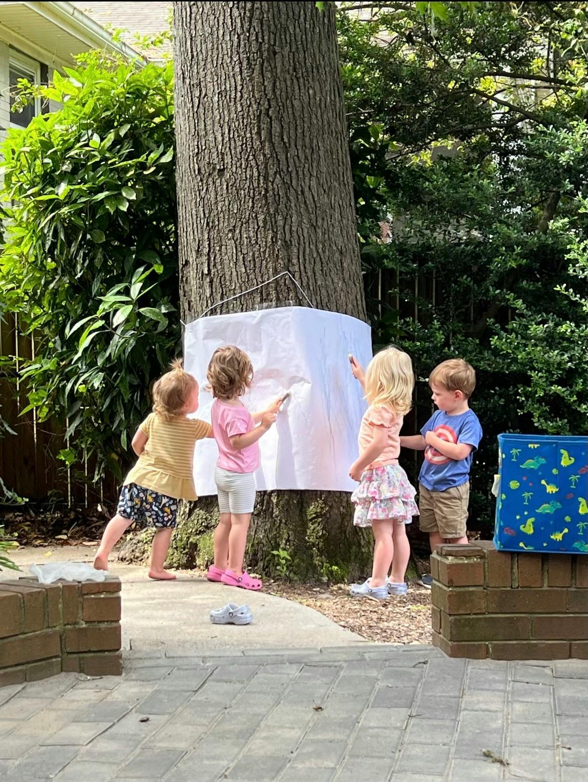 Four children painting on a large sheet of paper attached to a tree