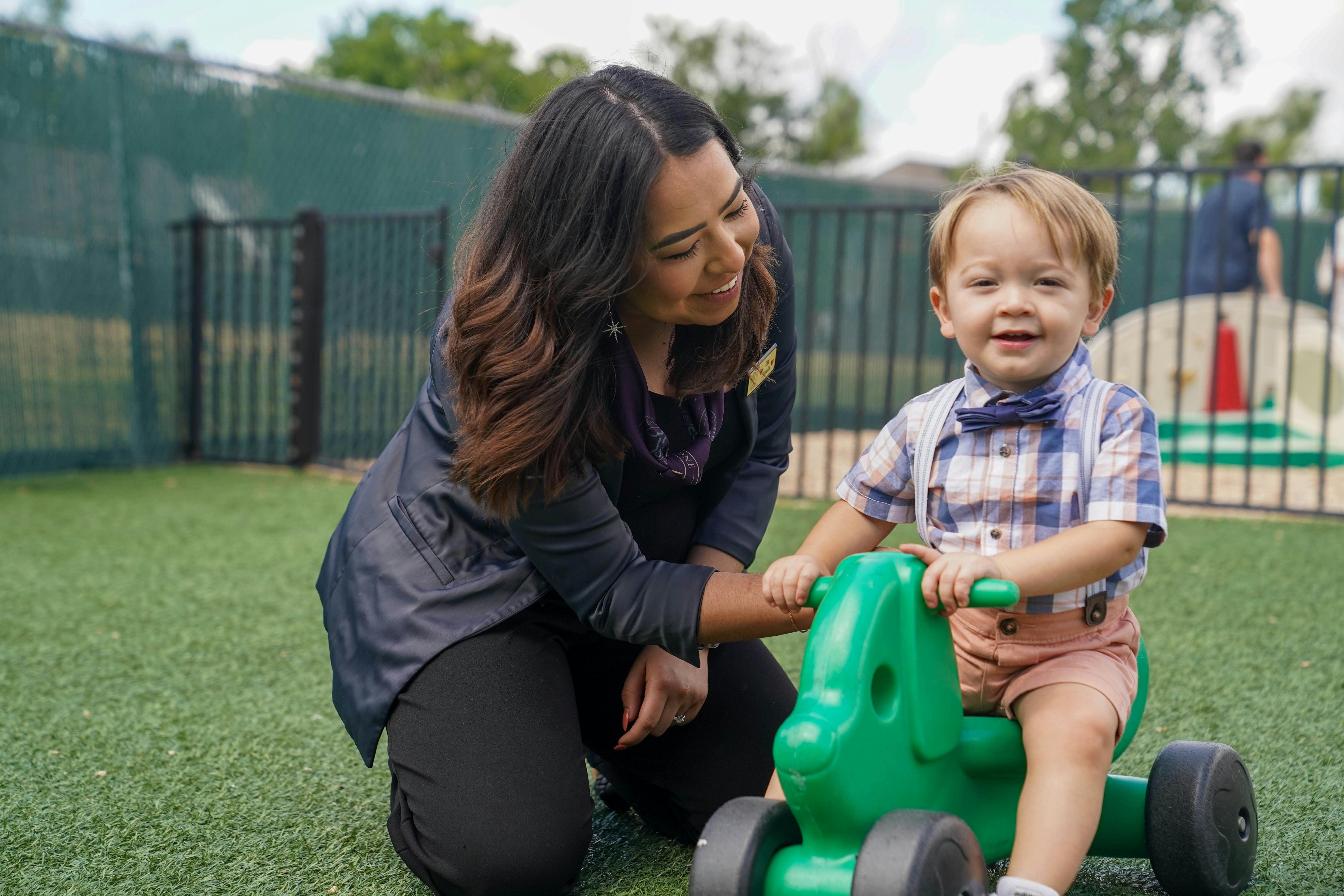 Adult kneeling next to a young child on a green ride-on toy on artificial grass