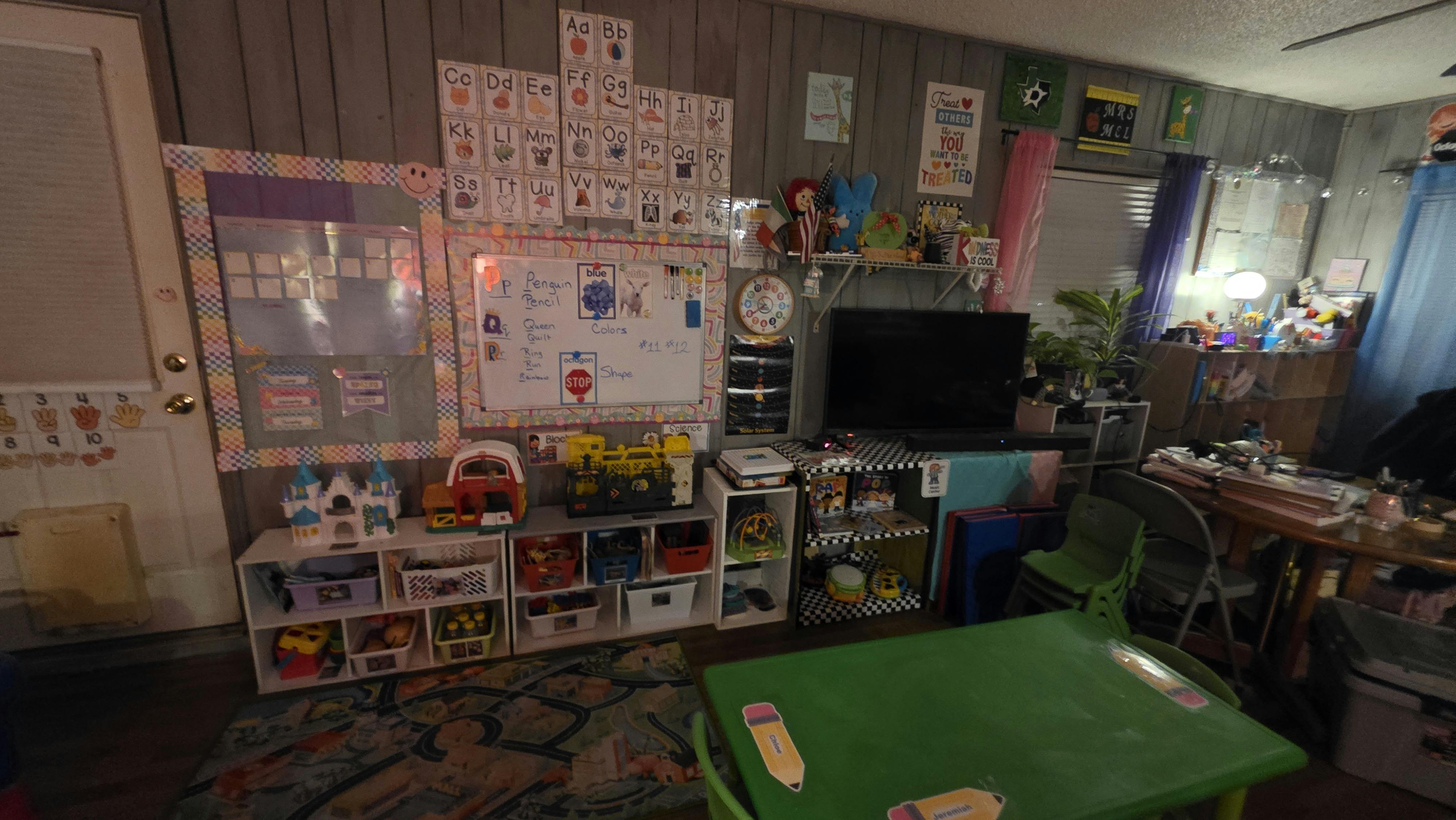 A colorful classroom with educational charts, a whiteboard, toy shelves, and a green children's table