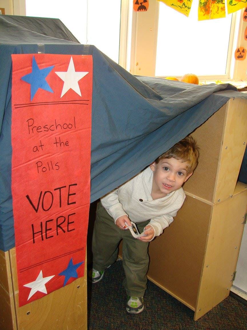 Child peeking out from under a blue sheet covering cardboard boxes, next to a "VOTE HERE" sign