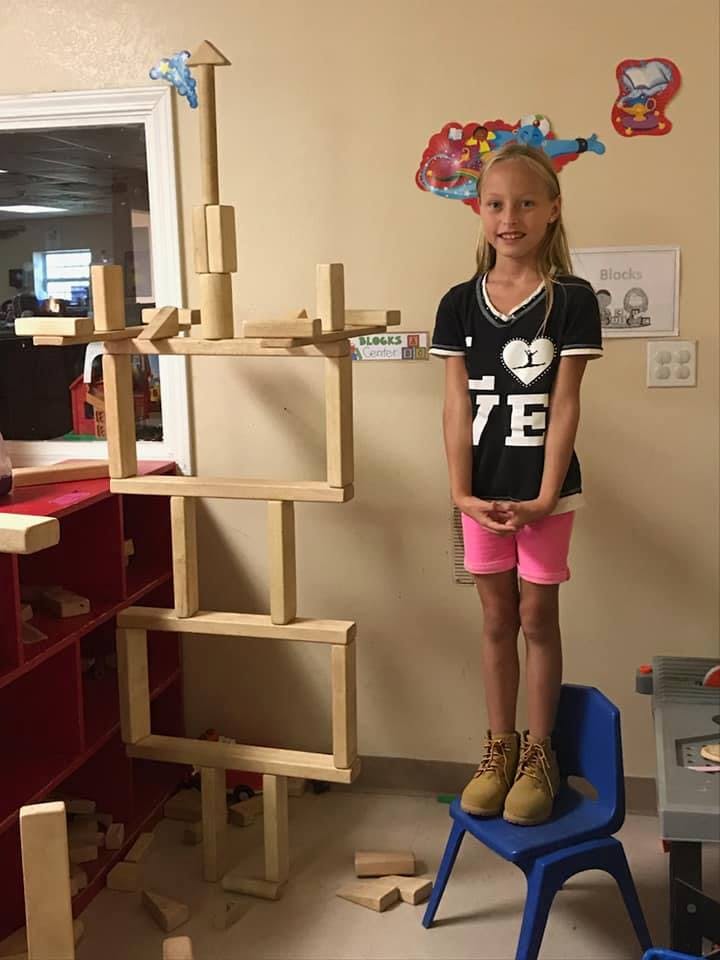Child standing on a chair next to a tall structure made of wooden blocks
