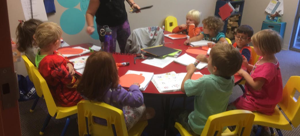 Children sitting at a table with papers and a person standing nearby
