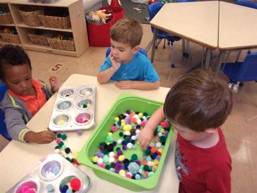 Three children playing with colorful pom-poms and muffin tins in a classroom