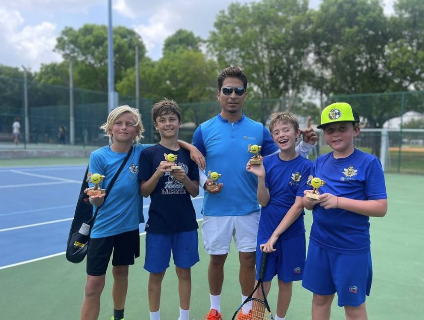 Adult and four children holding trophies on a tennis court