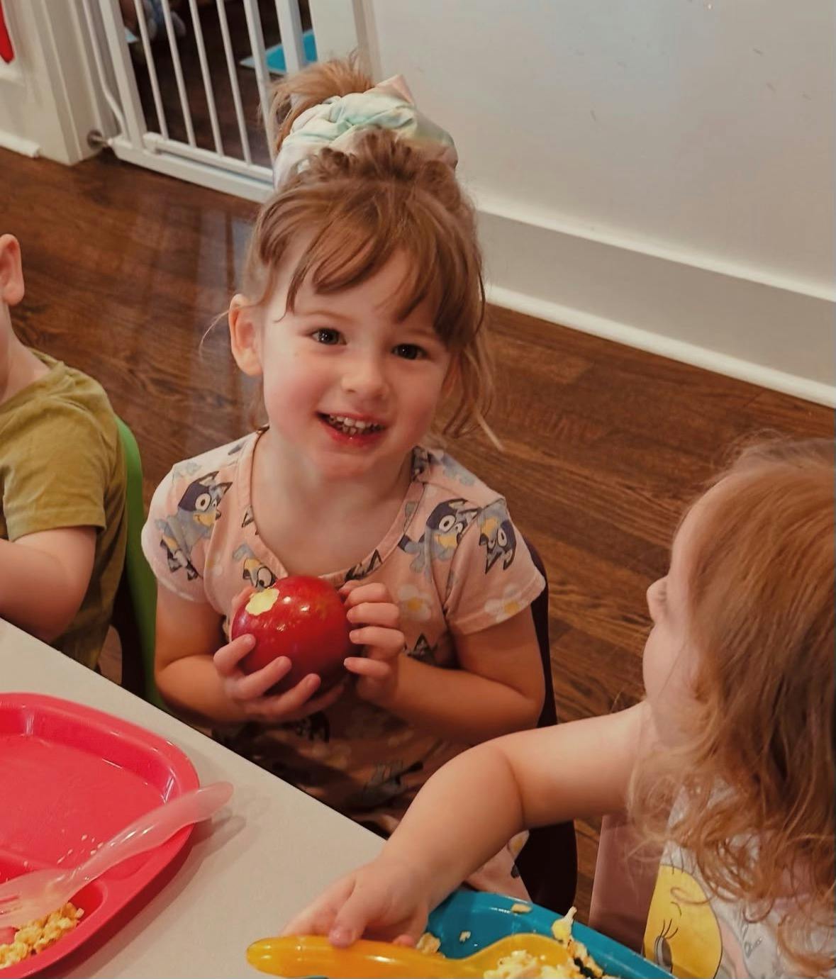 Child holding a red apple at a table with other children