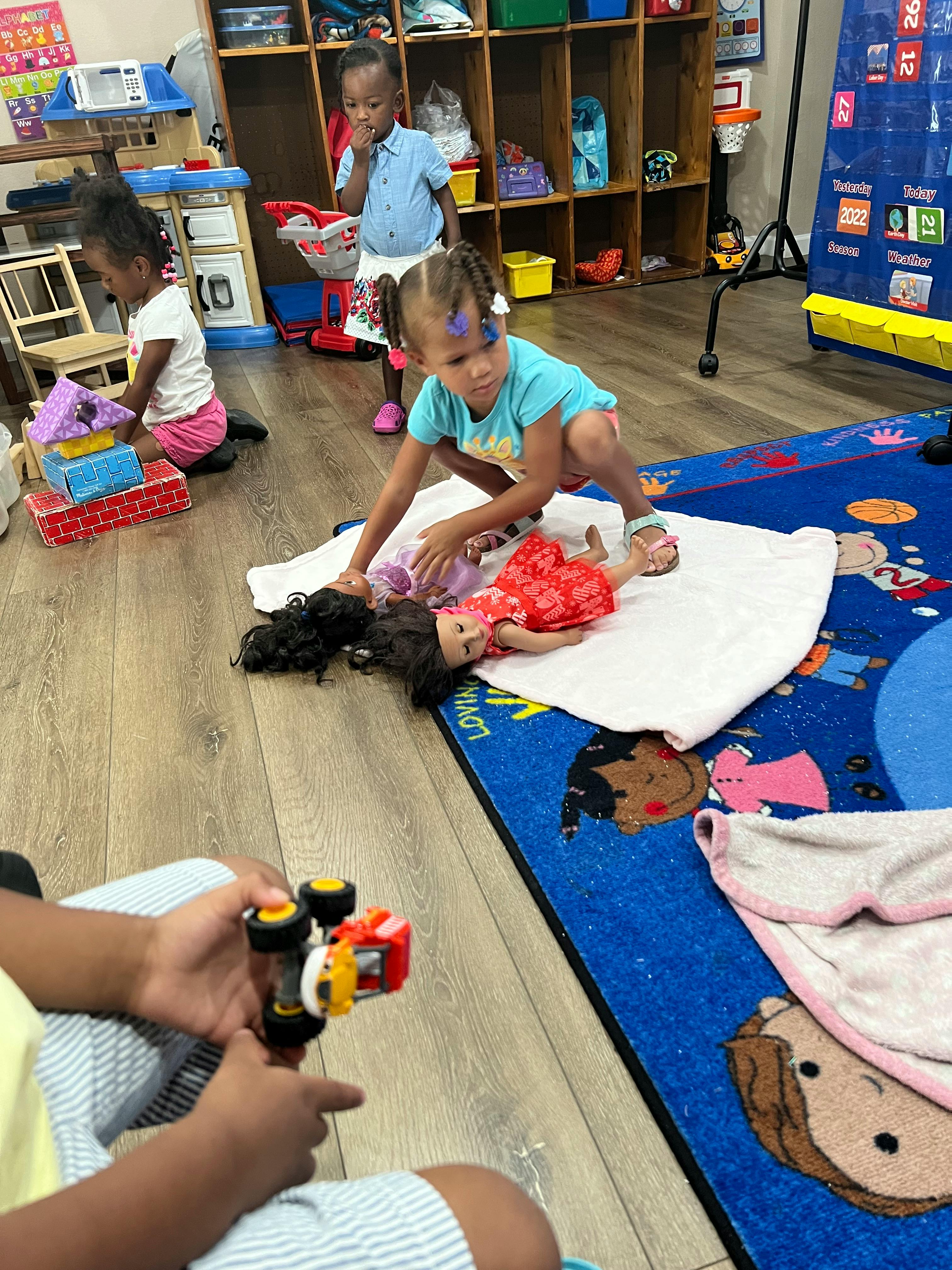 Child playing with a doll on a rug, other children playing in the background