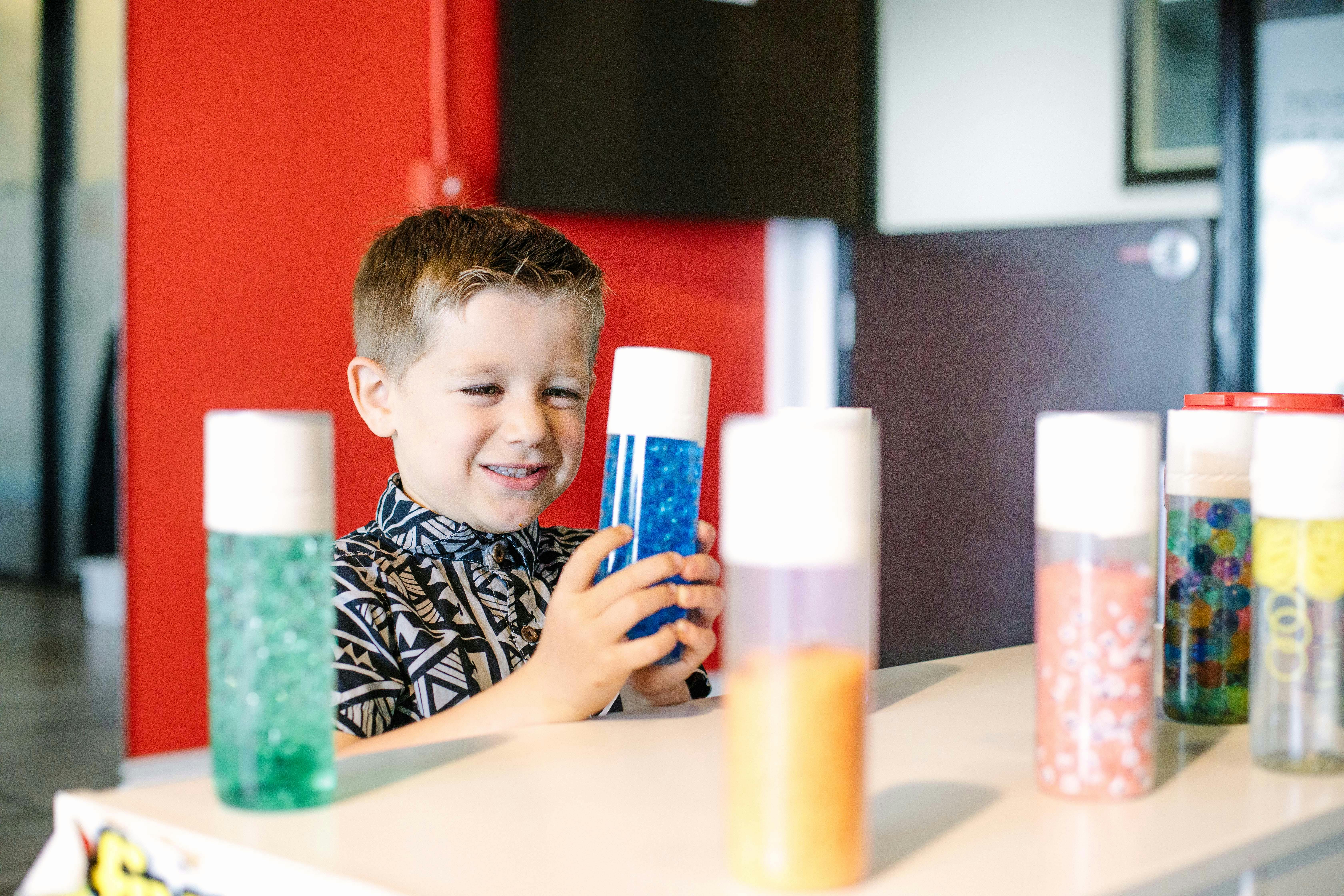 Child holding a blue sensory bottle at a table with other sensory bottles