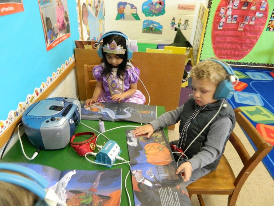 Two children wearing headphones and reading books at a table