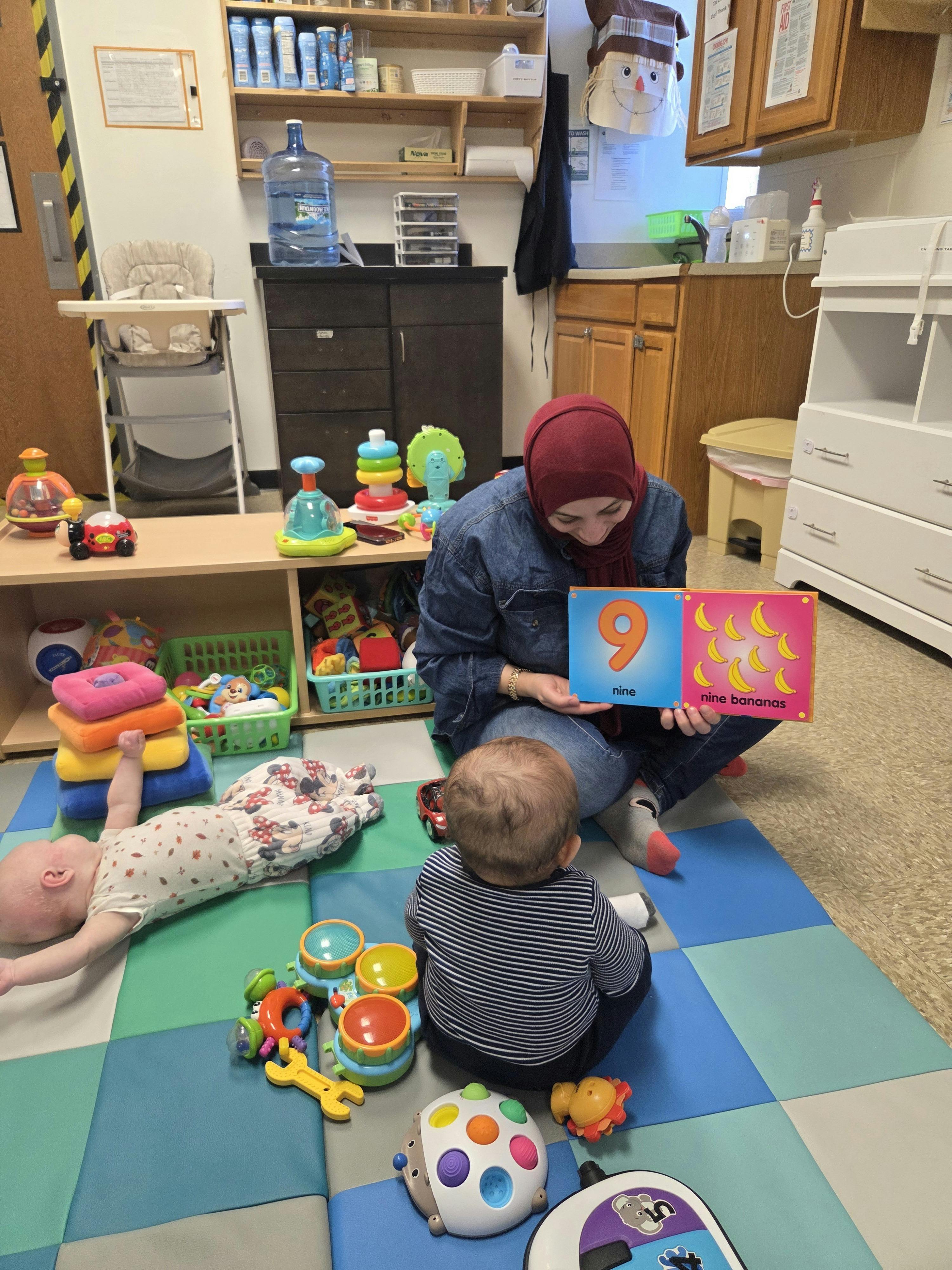 Person in a headscarf holding up a book for two children on a play mat