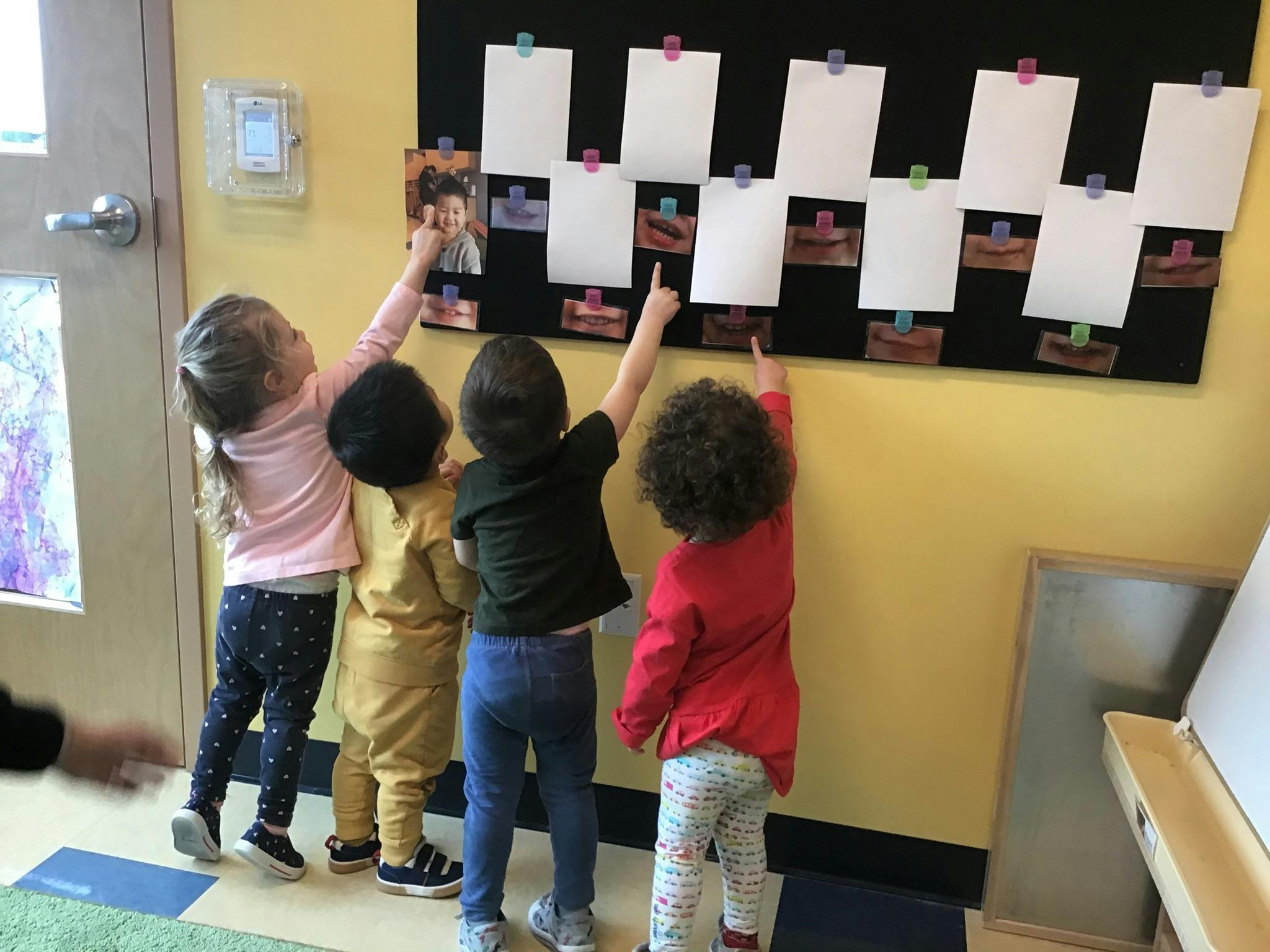Four children pointing at a bulletin board with papers in a classroom