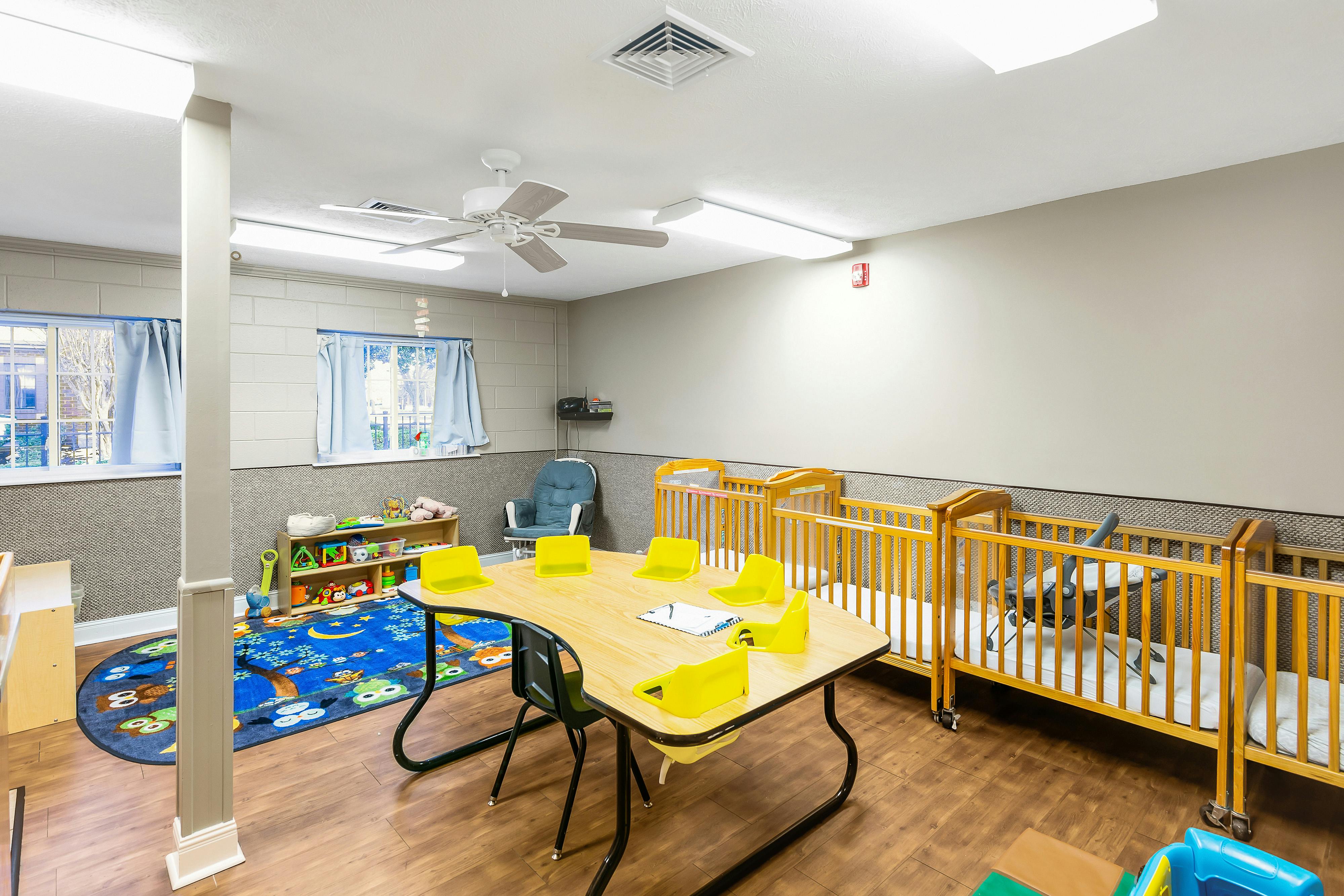 A classroom with cribs, a child-sized table with yellow chairs, and play items