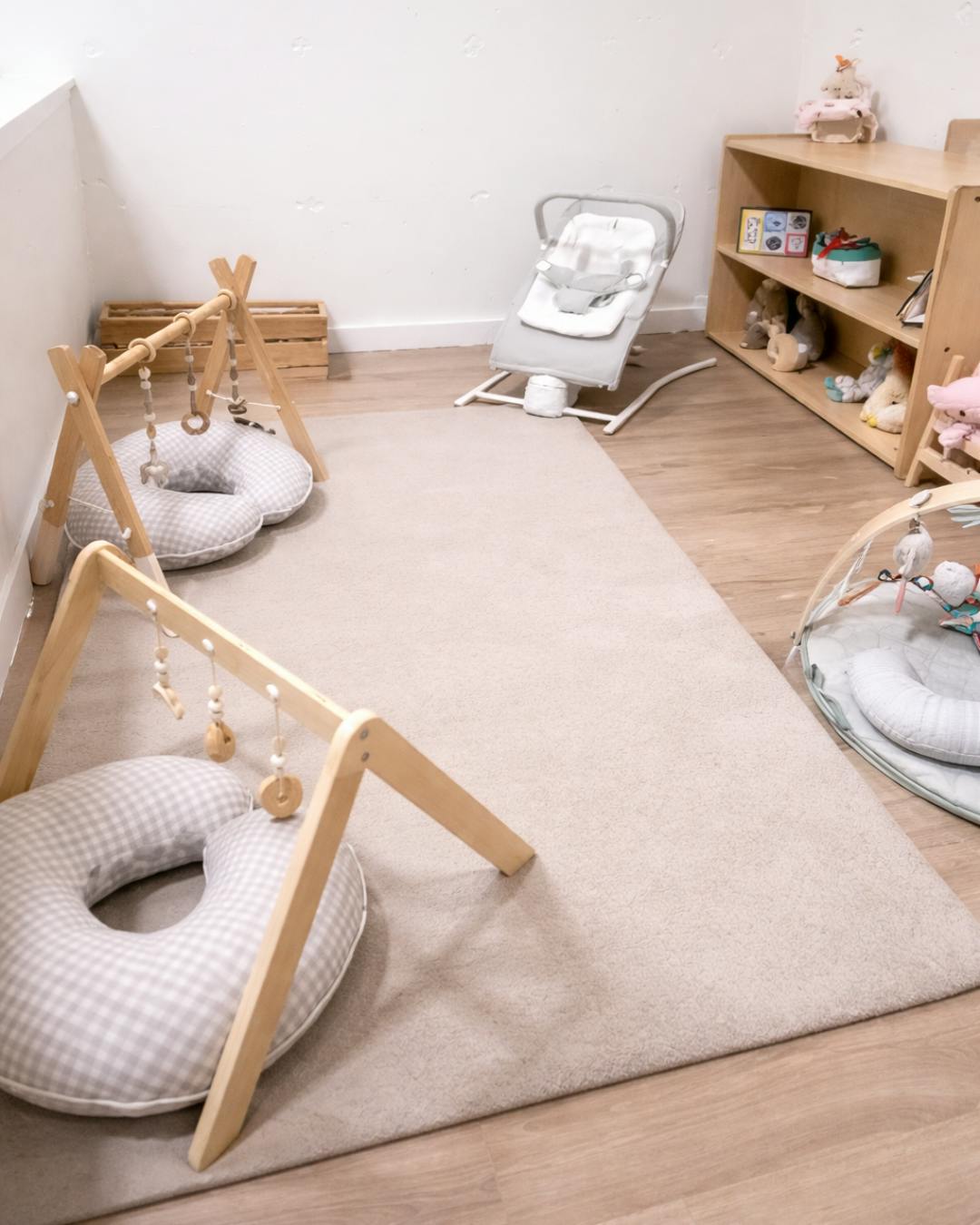 Baby play area with two play gyms, a bouncer, and a wooden shelf with toys