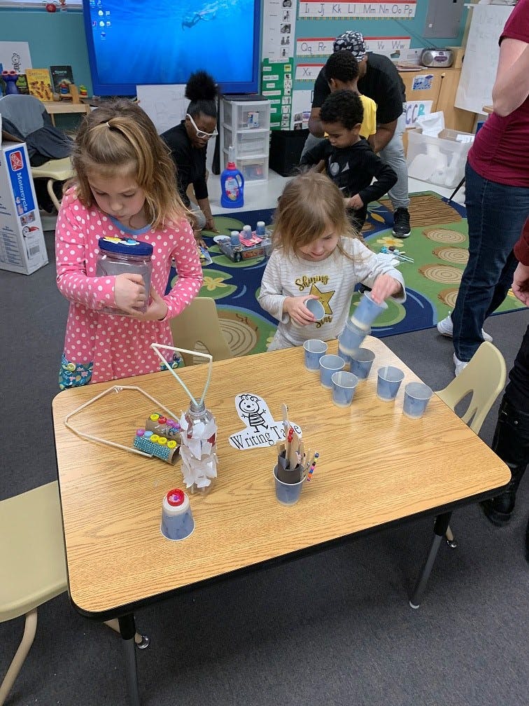 Two children playing with cups and craft materials at a table in a classroom