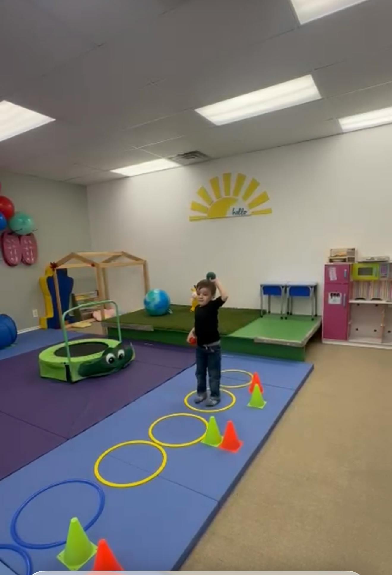 Child standing on blue mat with hoops and cones, holding a ball