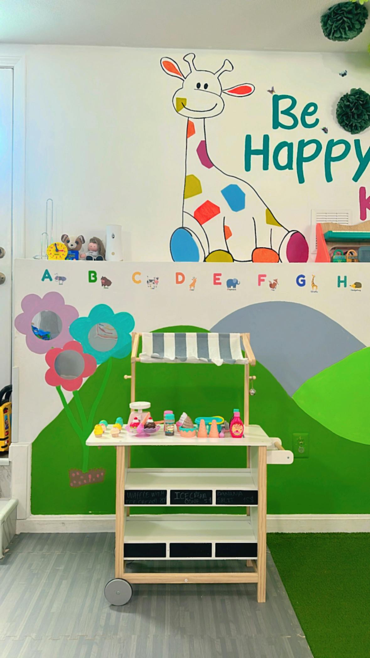 Play kitchen with toy food in a room with a giraffe mural and "Be Happy" text