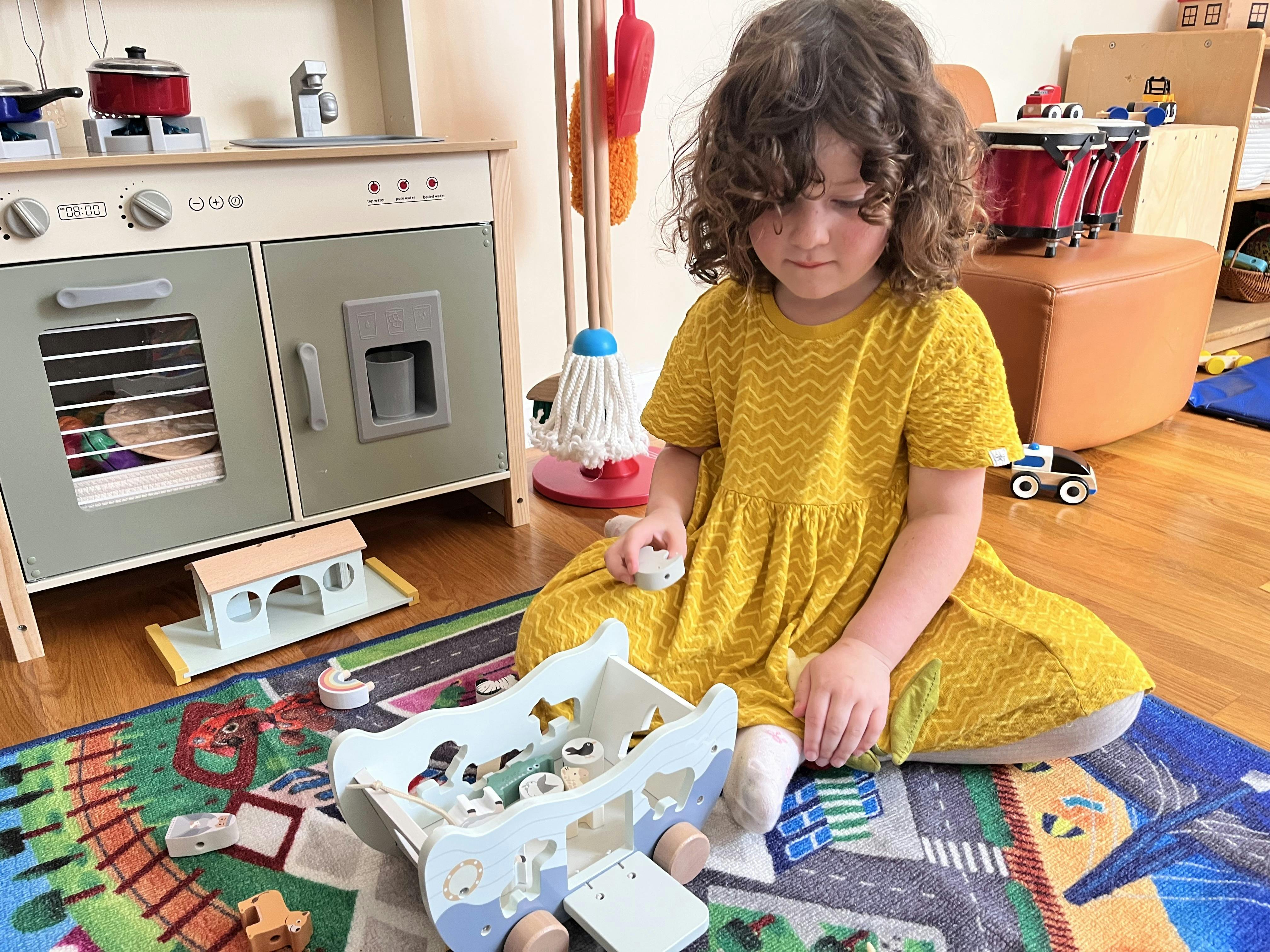 Child in a yellow dress playing with a wooden toy boat on a play mat