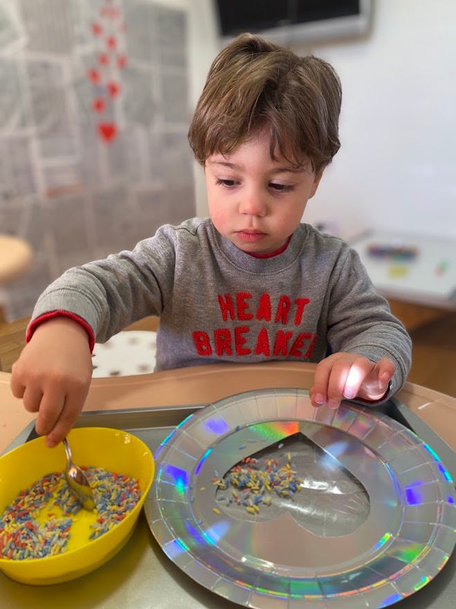 Child using a spoon to move colorful rice from a bowl to a heart-shaped plate