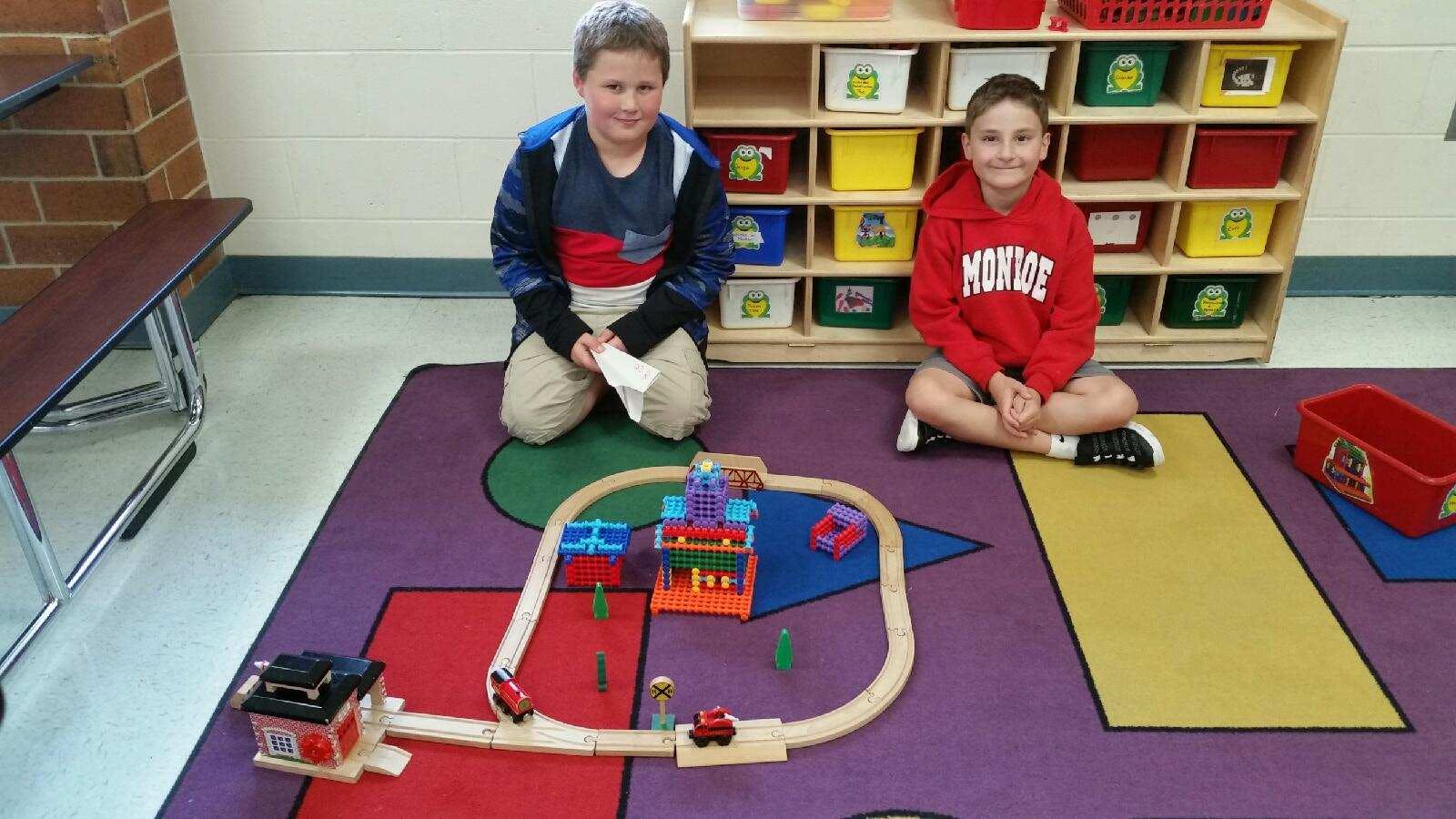 Two children kneeling on a rug with a toy train set