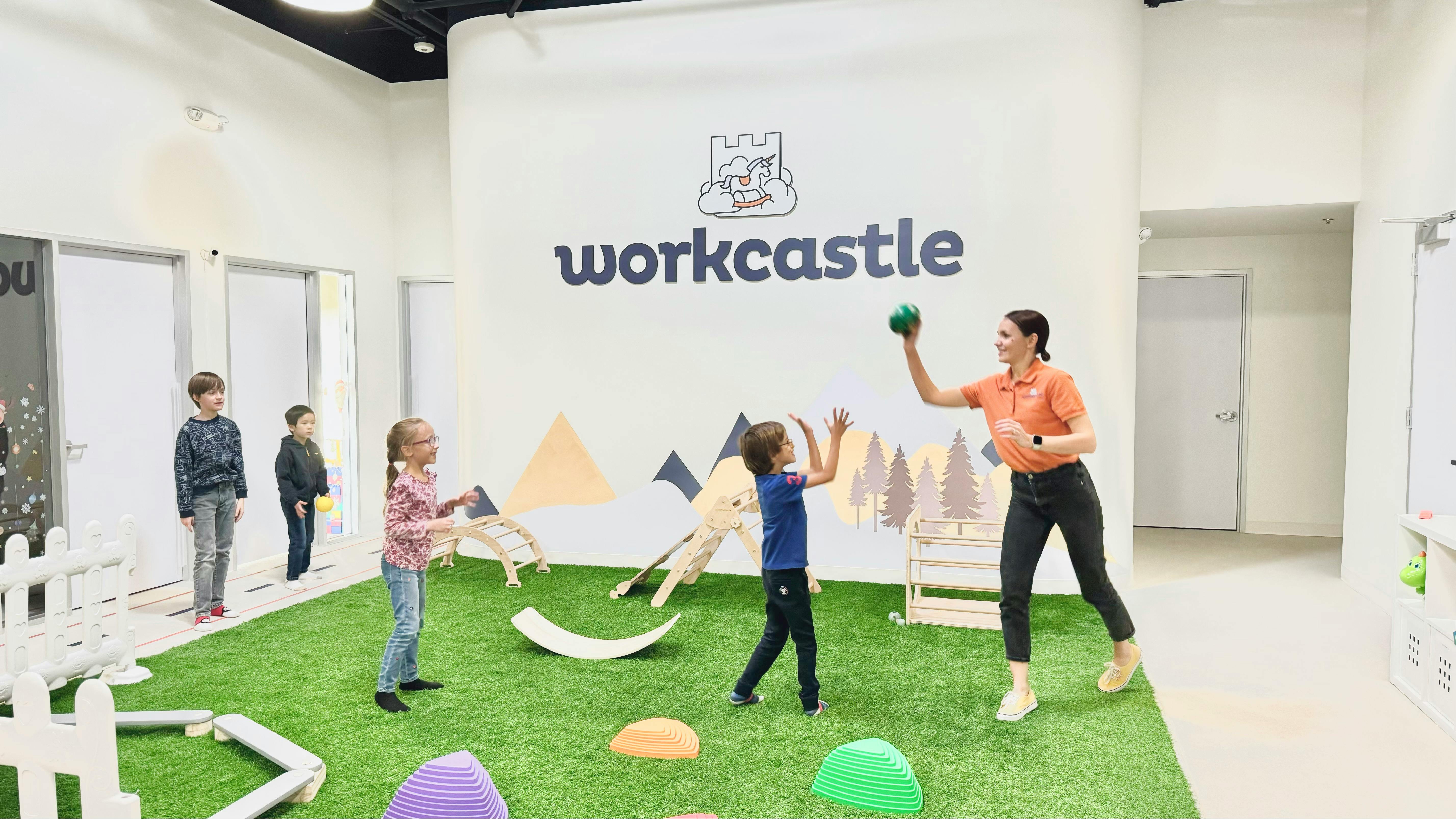 Adult and children playing with a ball on a green turf area with "workcastle" on the wall
