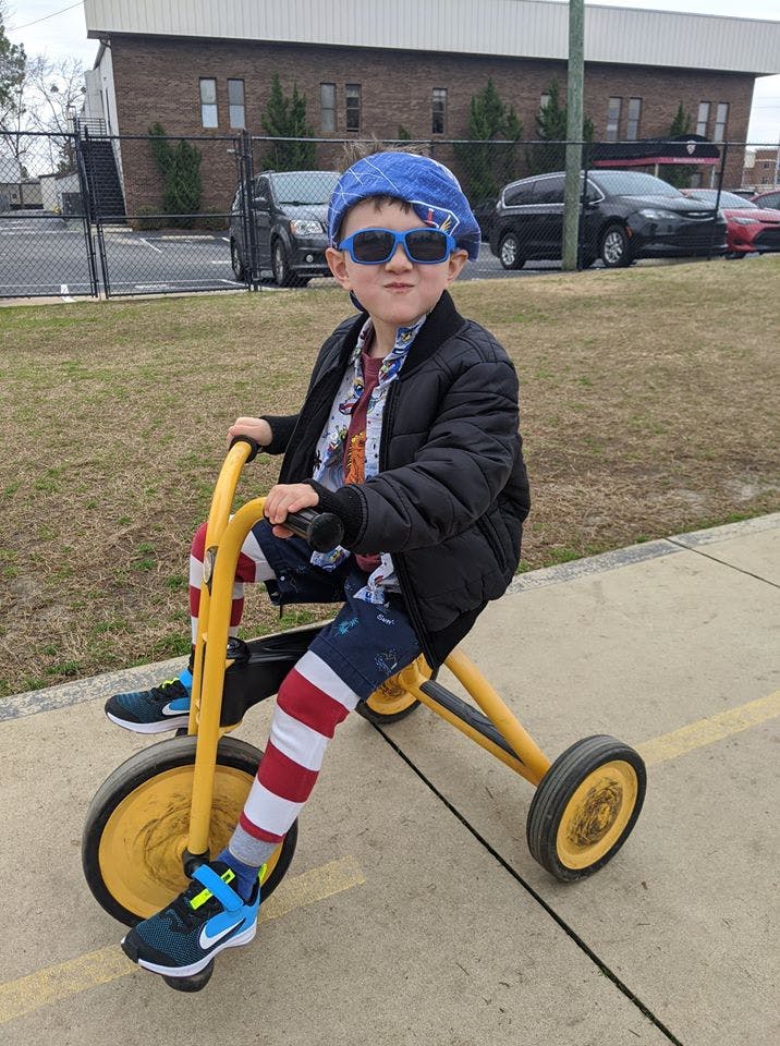 Child in sunglasses and blue hat riding a yellow tricycle