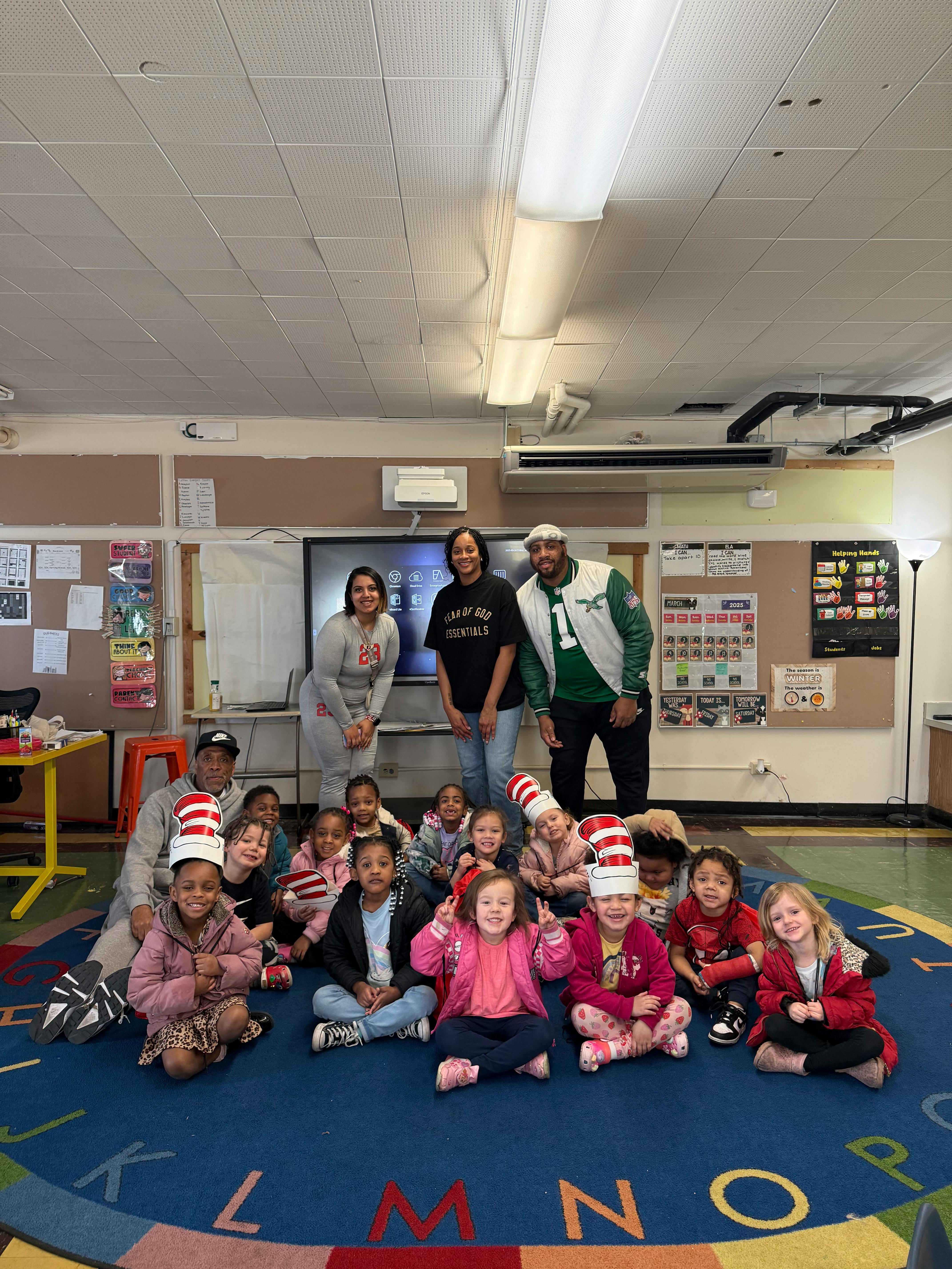 Adults and children sitting on a rug in a classroom, some wearing striped hats