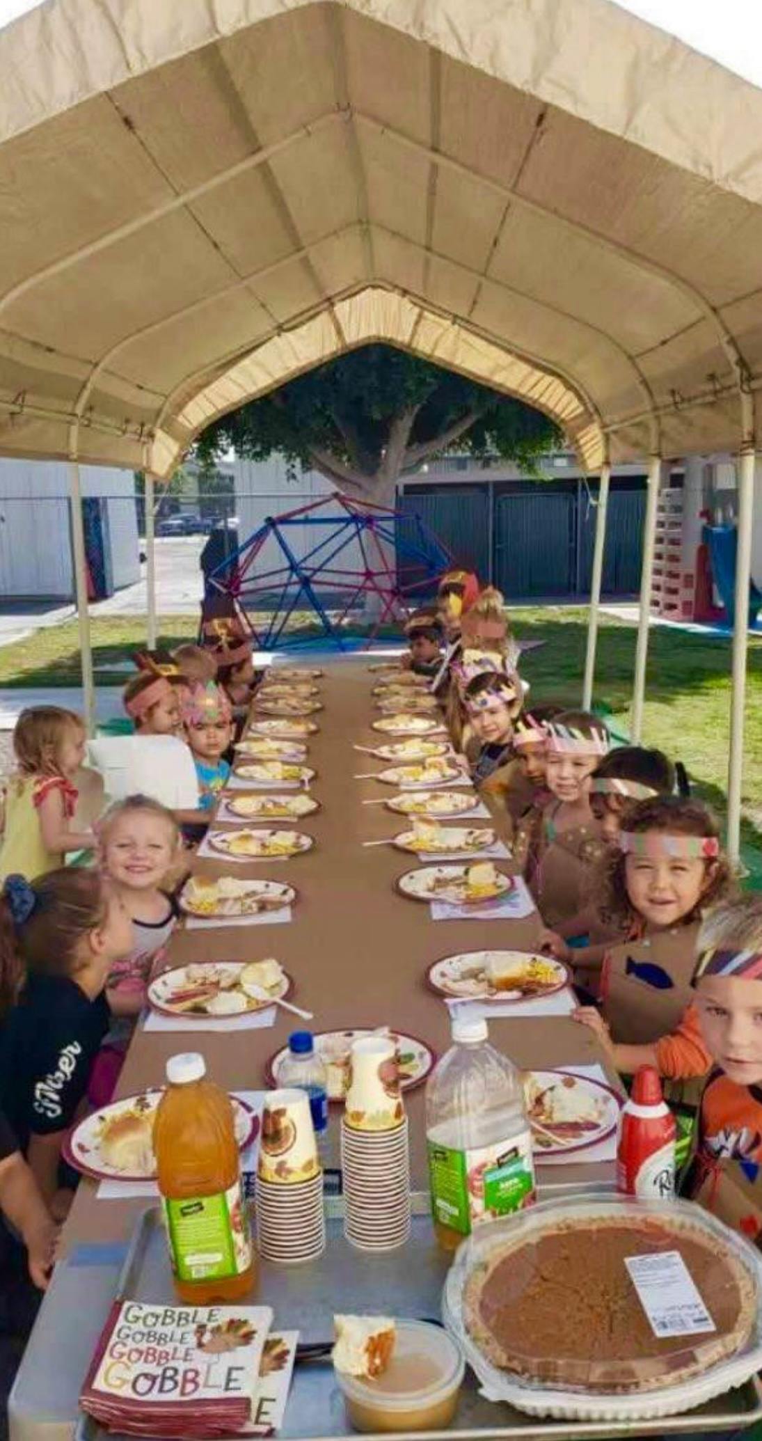 Children sitting at a long table with plates of food under a canopy