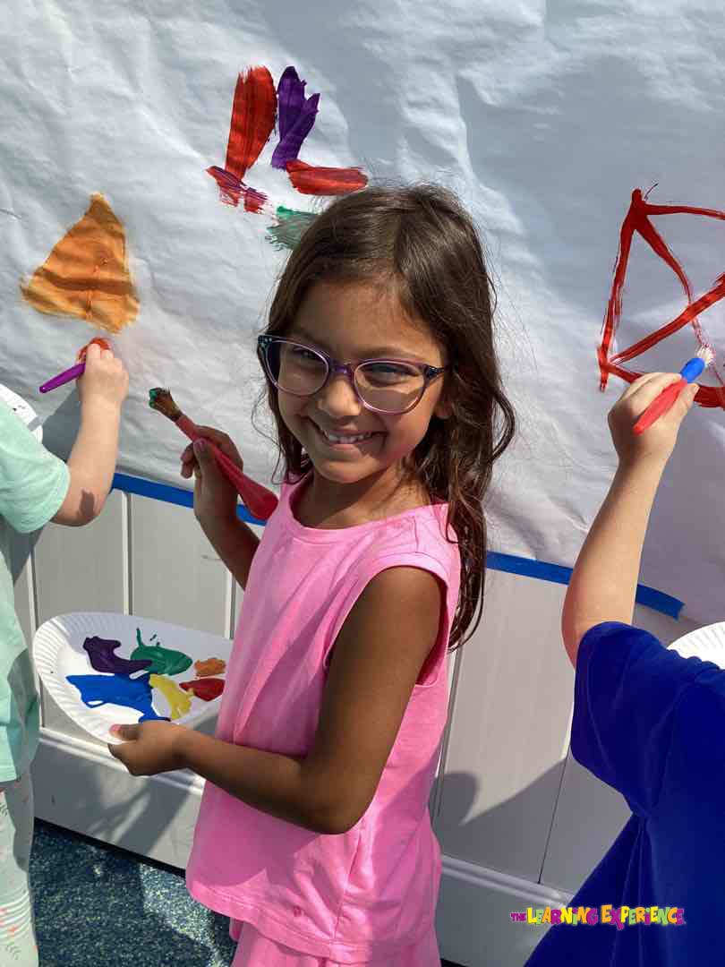 Child in glasses holding a paint palette and brush, painting on a white surface