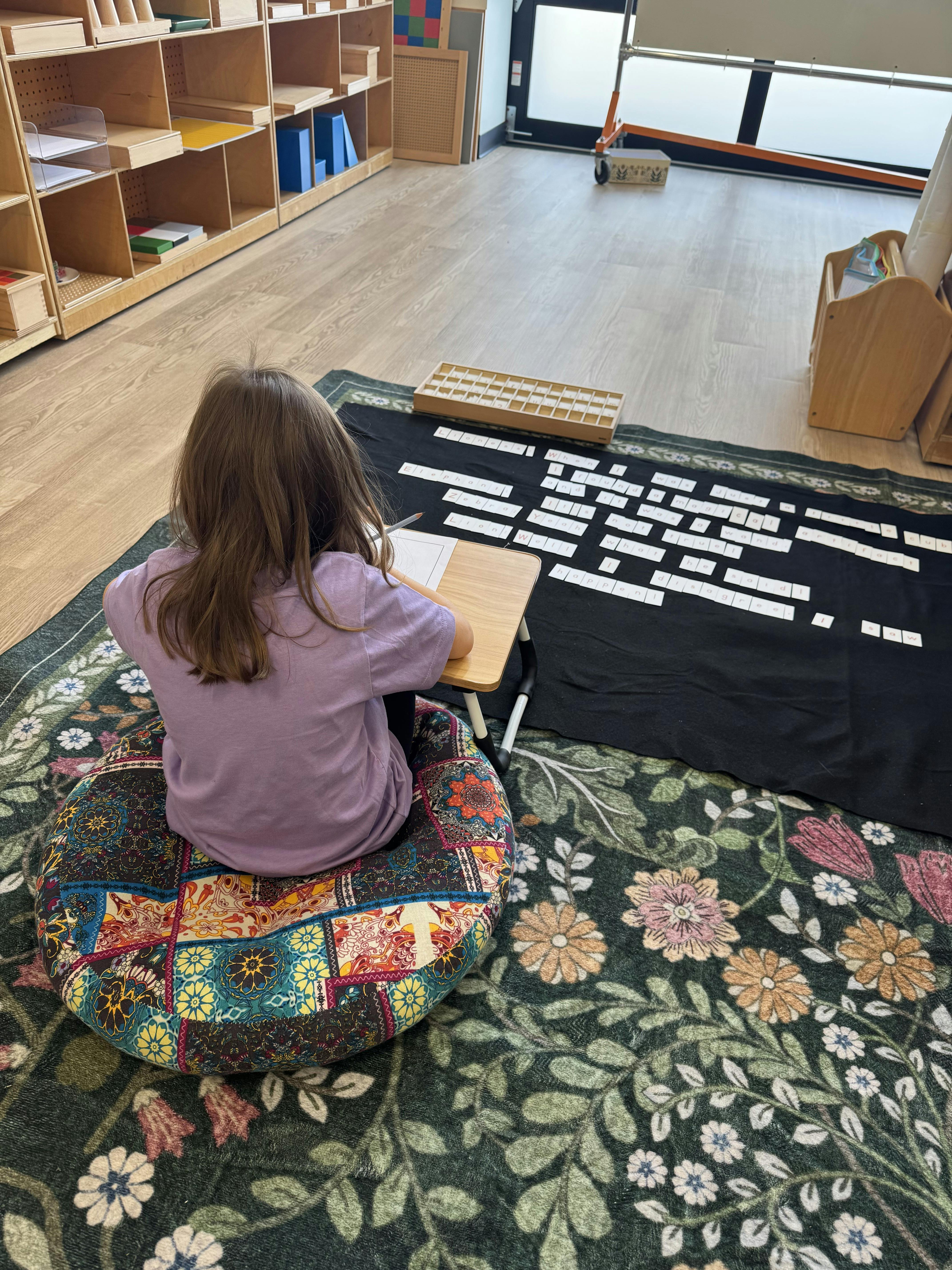 Child sitting on a floor cushion using a small desk to write near word cards