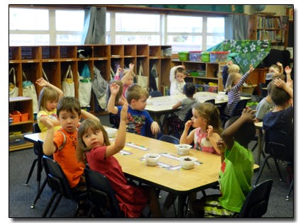 Children sitting at tables in a classroom with some raising their hands