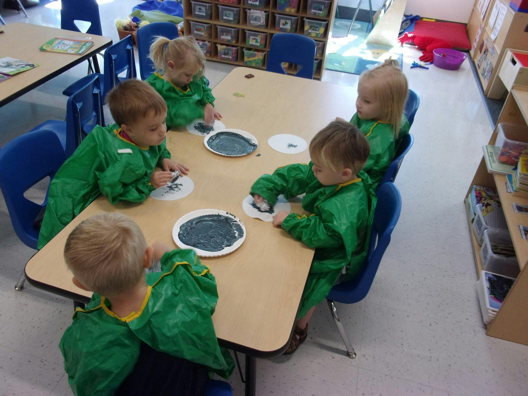 Children in green smocks painting at a table in a classroom