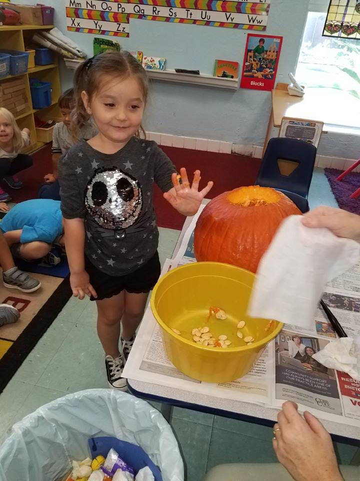 Child with pumpkin seeds on fingers next to a pumpkin and yellow bowl