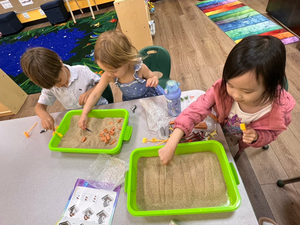 Three children playing with sand in green trays at a table