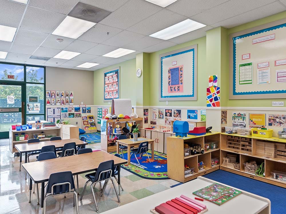 Empty classroom with small tables and chairs, shelves with toys, and posters