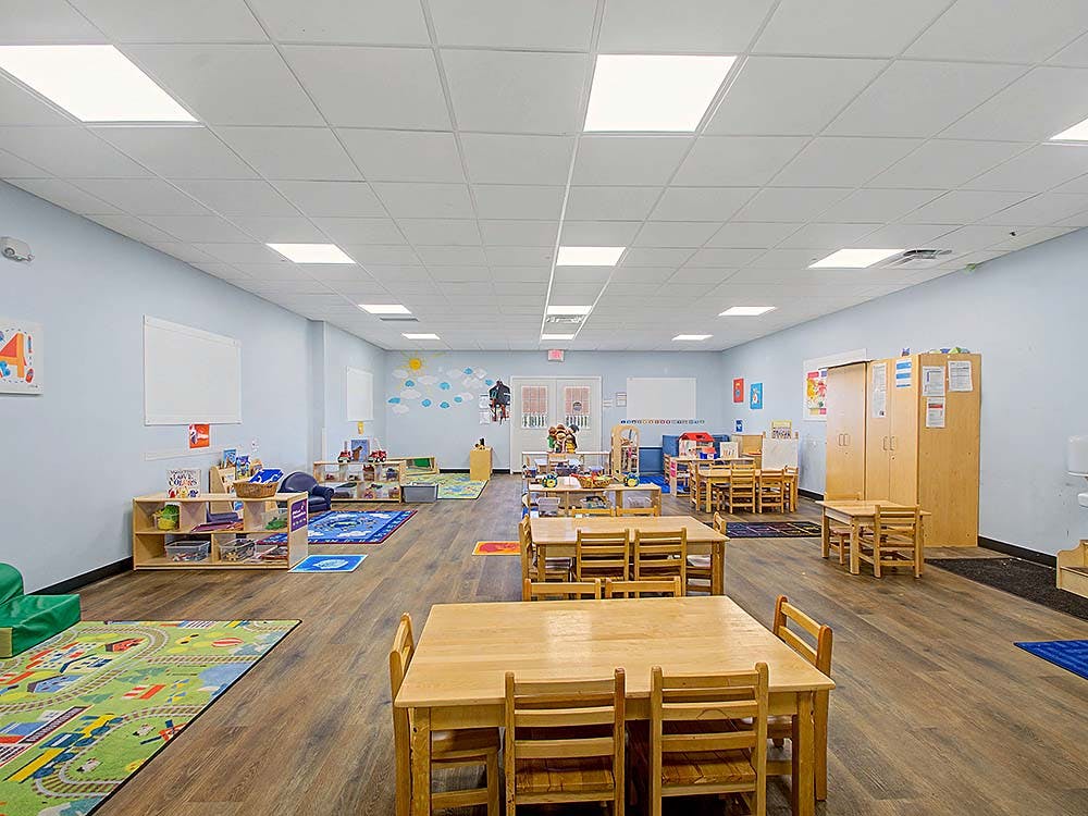 Empty classroom with small wooden tables and chairs, play rugs, and shelving
