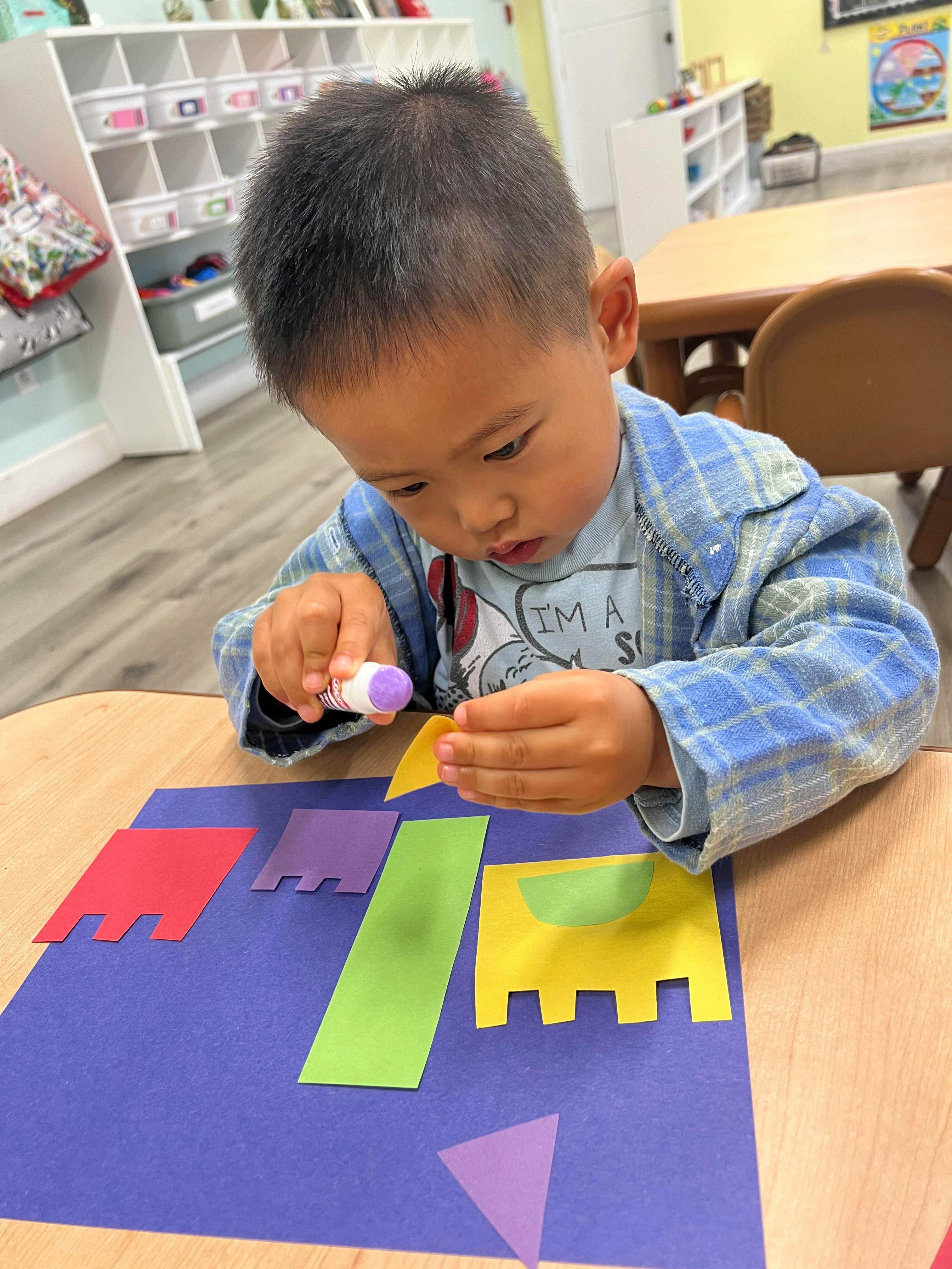 Child gluing paper shapes onto a sheet of paper at a table