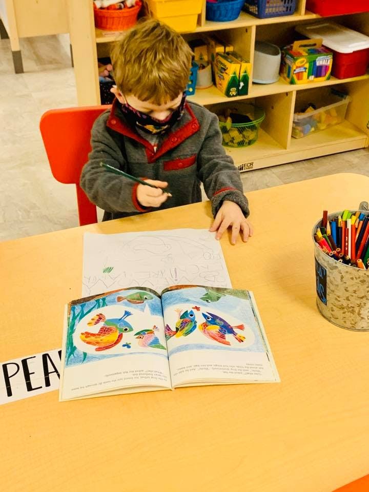 Child in a mask drawing at a table with an open book