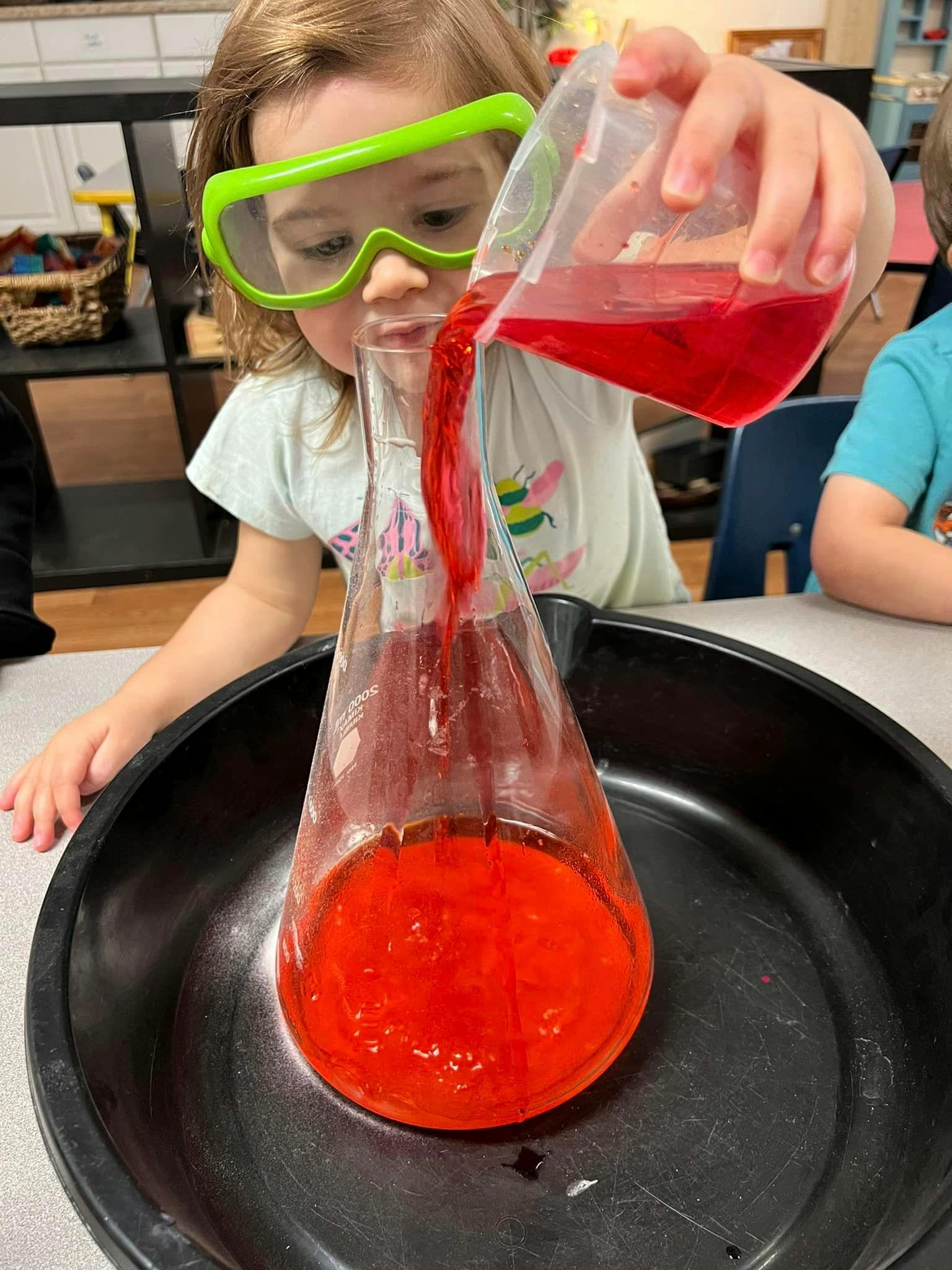 Child in safety goggles pouring red liquid into a flask